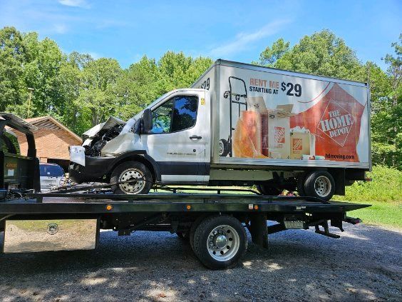 White box truck on a flatbed trailer with front-end damage, parked outdoors near trees and a house.