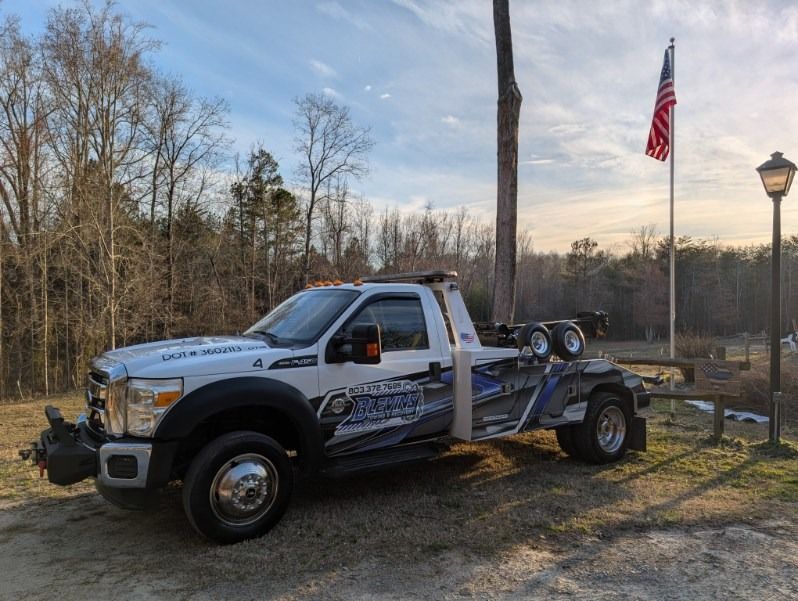 White tow truck on roadside at sunset, with American flag and trees in the background.