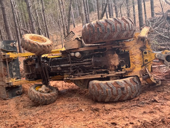 Upside-down yellow tractor in a muddy forest, its large tires exposed after a rollover