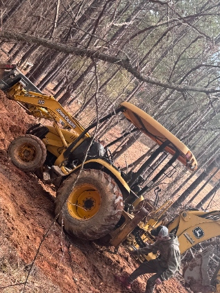 Yellow construction loader and excavator on a muddy, wooded hillside with a worker nearby