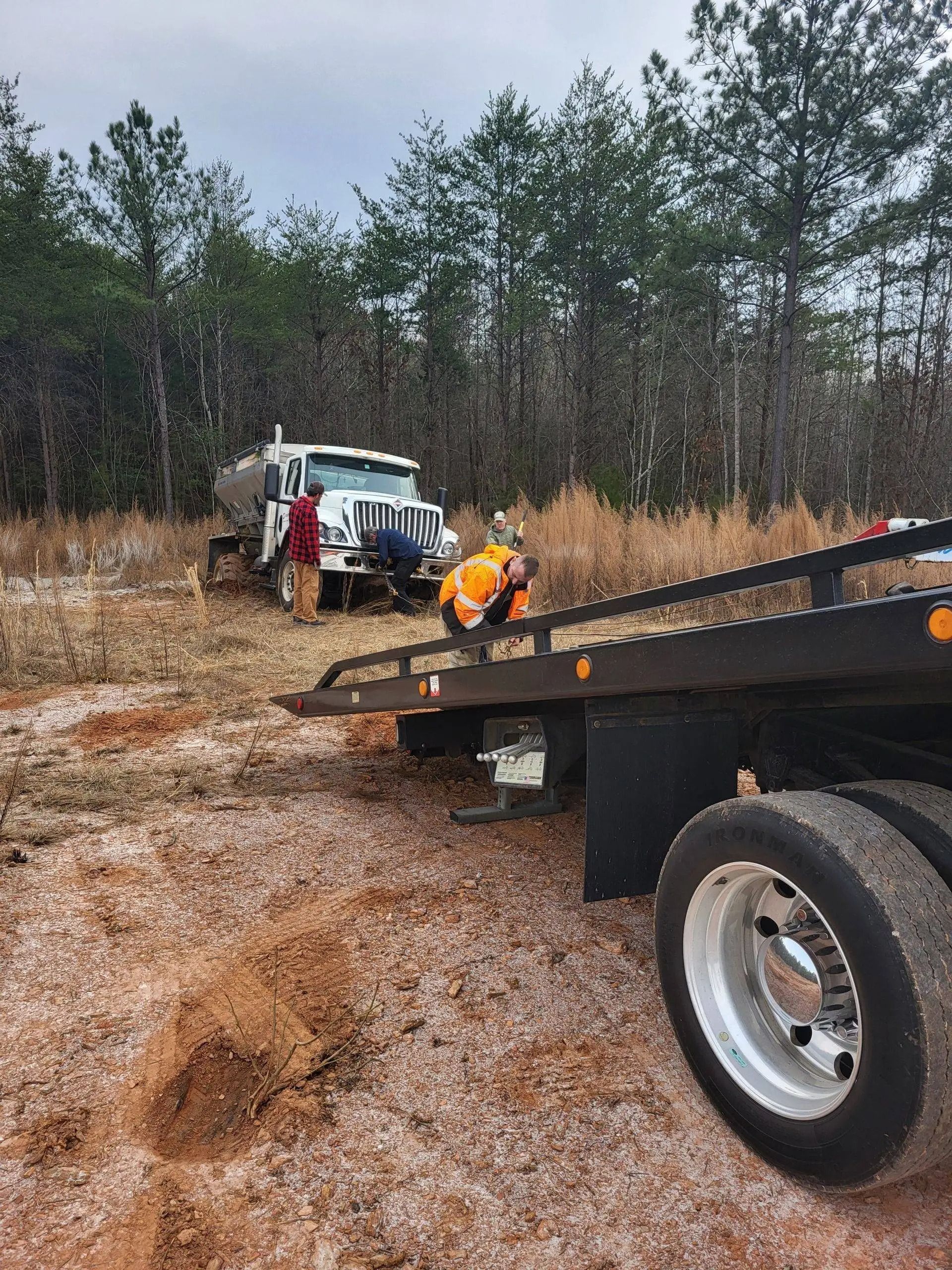 Tow truck pulling a flatbed trailer on a snowy dirt road with workers and pine trees in the background
