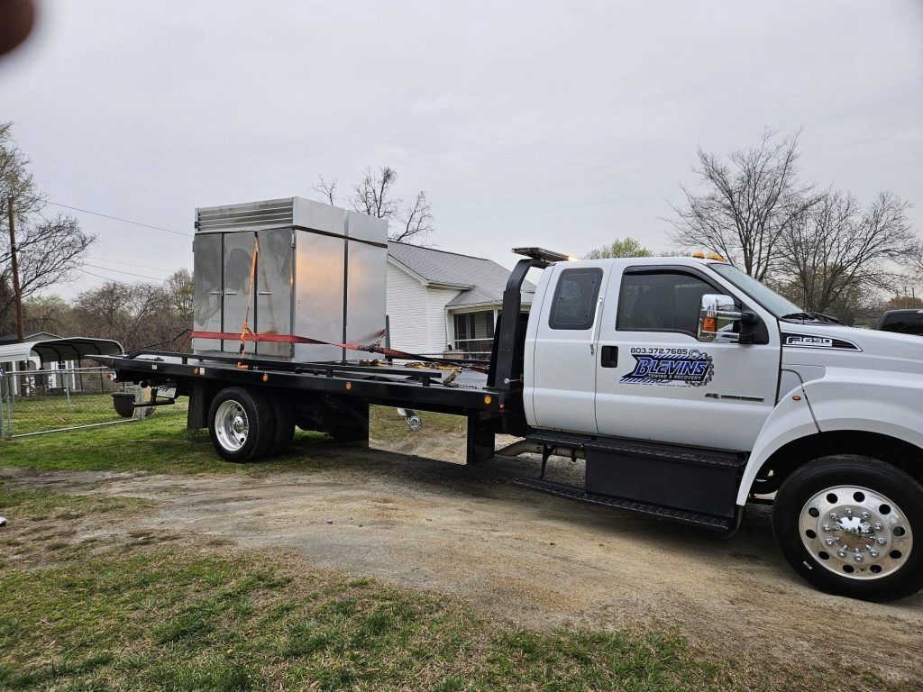 White flatbed truck carrying a large metal container on a gravel lot under an overcast sky