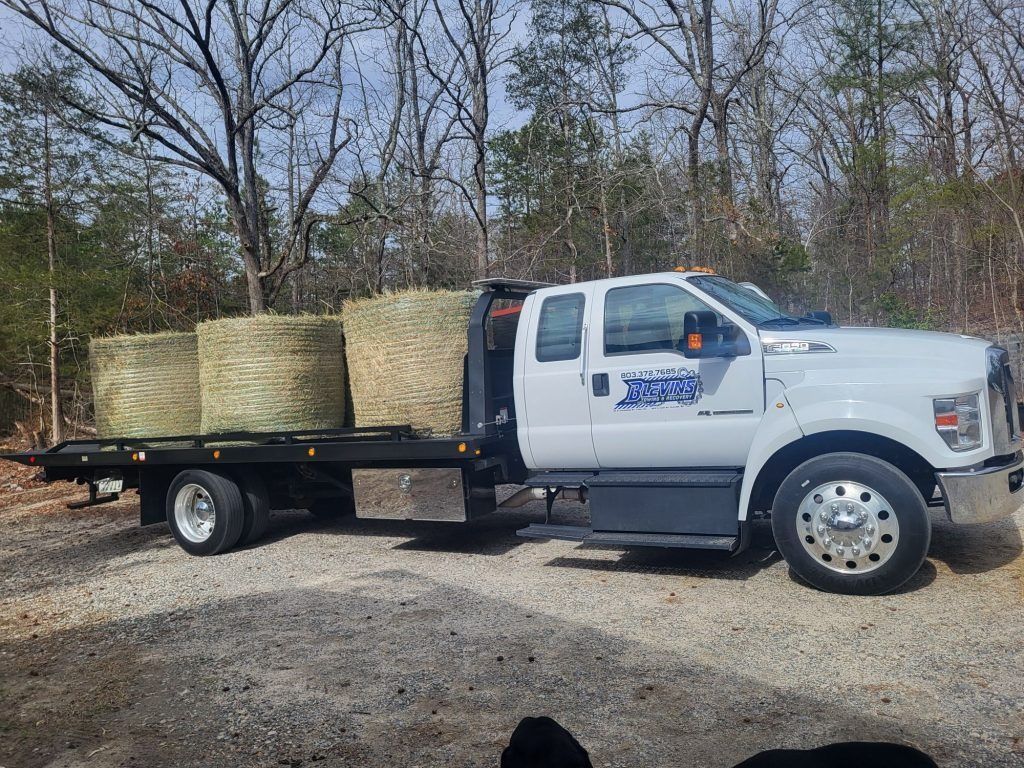 White pickup truck carrying large hay bales on a flatbed trailer in a gravel lot.