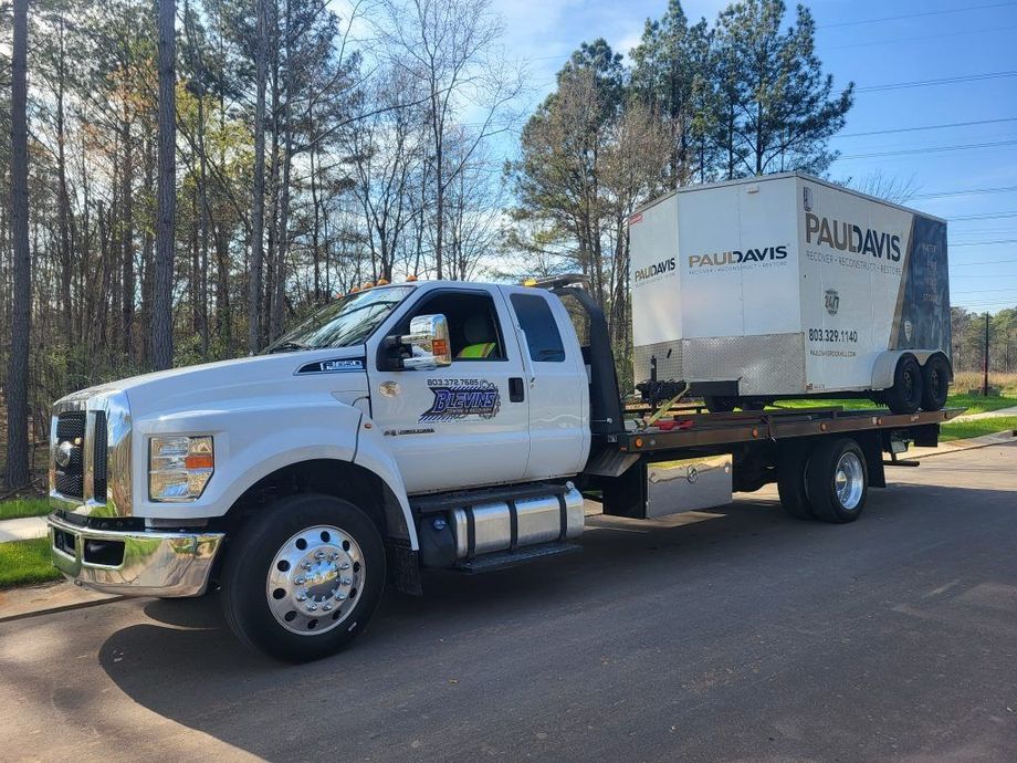 White flatbed truck parked on a road beside pine trees, carrying a large white container load