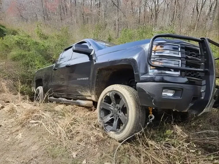 Black pickup truck stuck in a muddy ditch beside a wooded roadside
