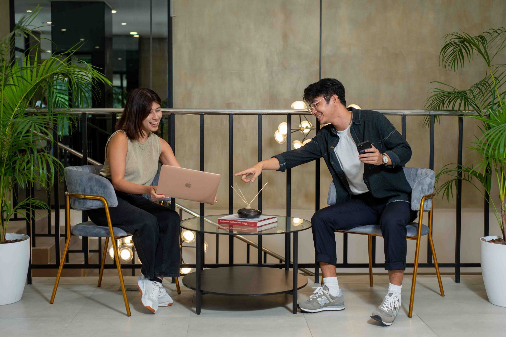 Two people sit in armchairs in a modern lounge, discussing work with a laptop and a small device on a glass coffee table.