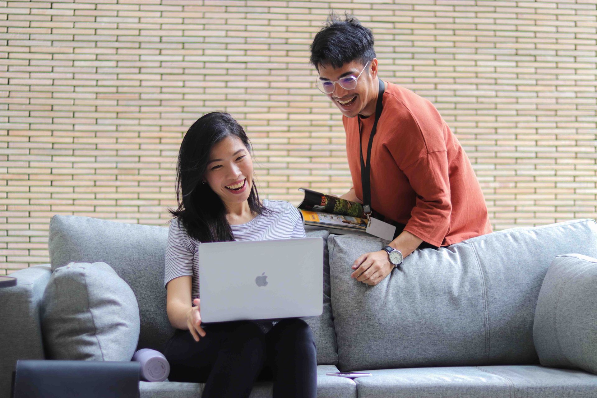 Two people smiling on a gray couch, looking at a laptop in a bright room.