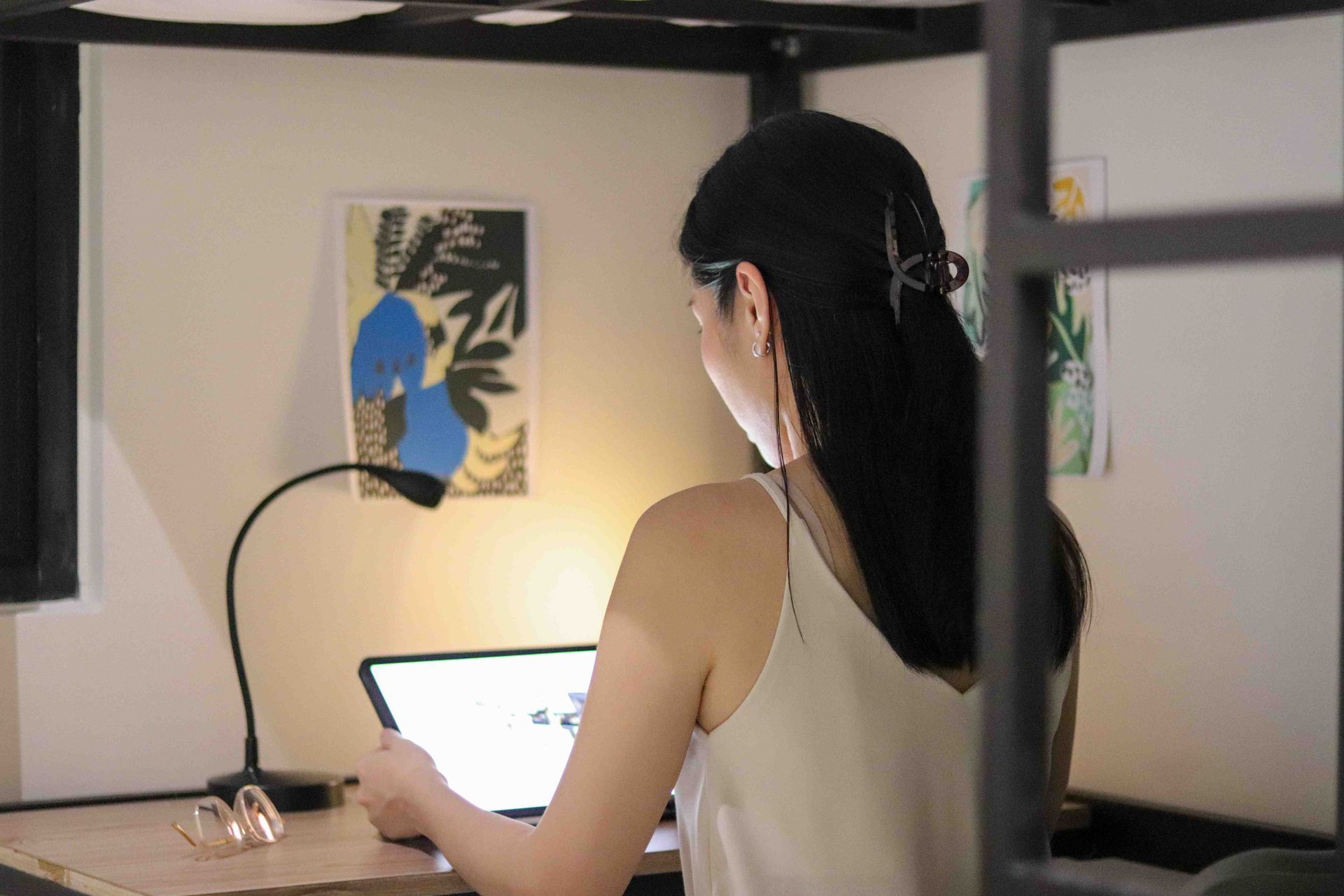 Woman working on a laptop at a desk, lit by a lamp, with art on the wall.