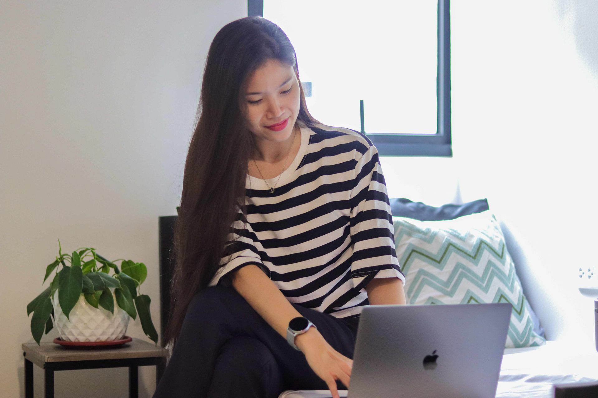 Woman sitting on a couch using a laptop beside a potted plant and patterned pillow