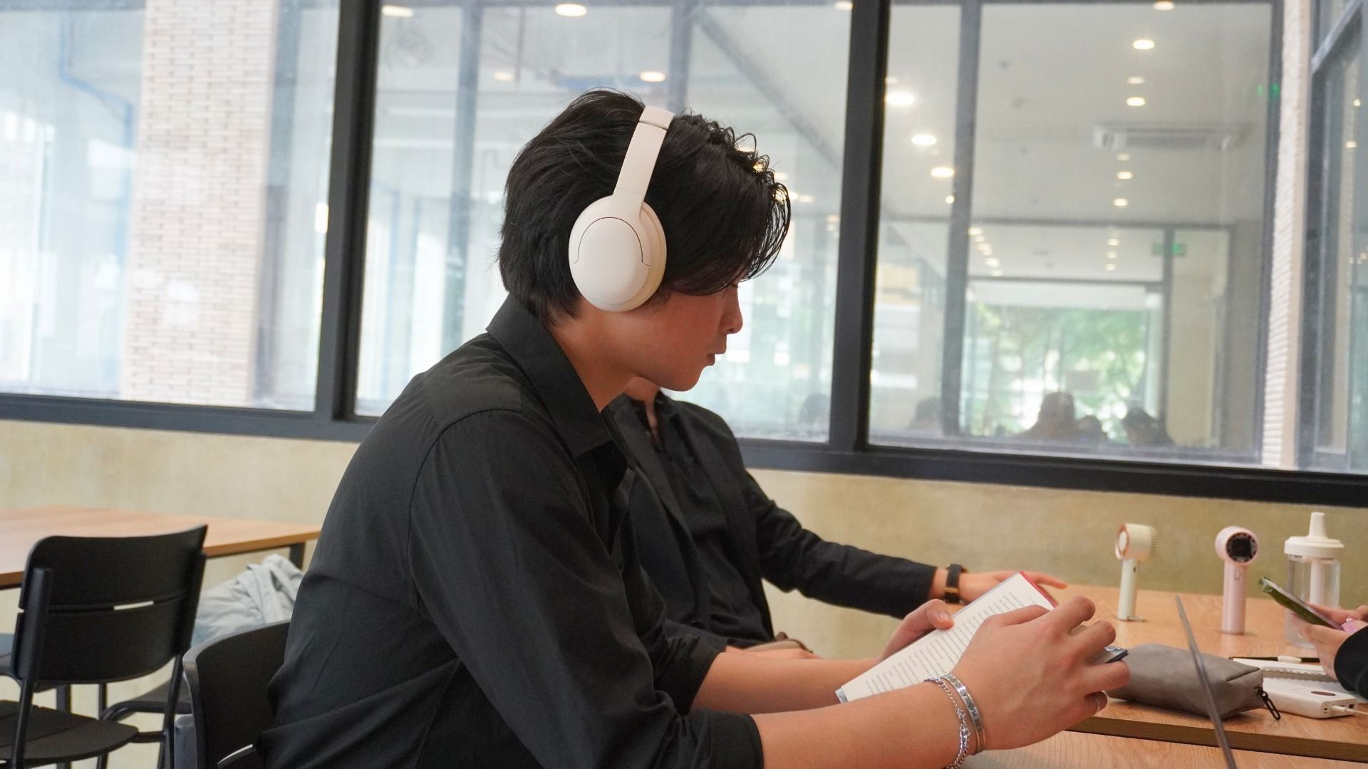 Man wearing white headphones, seated at a table, looking at a white book. Indoors near a window.