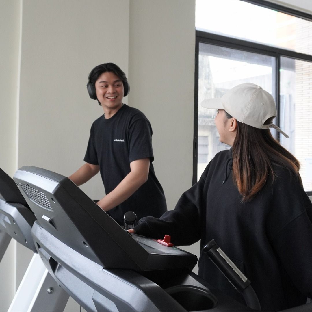 Two people on treadmills in a gym; one looks at the other, smiling.