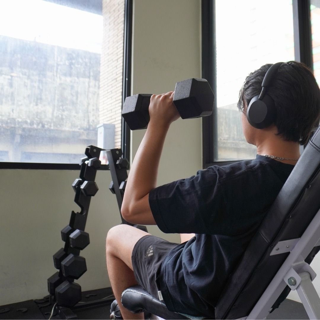 Person lifting a dumbbell while seated on a bench, wearing headphones in a bright room.