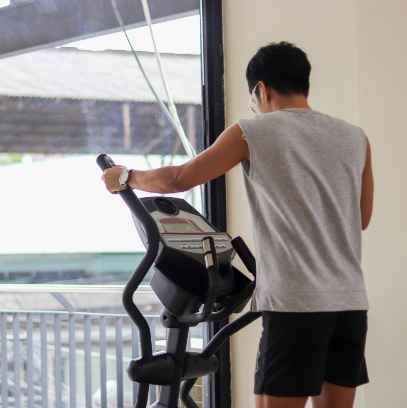 Man wearing gray tank top and black shorts exercising on an elliptical machine near a window.