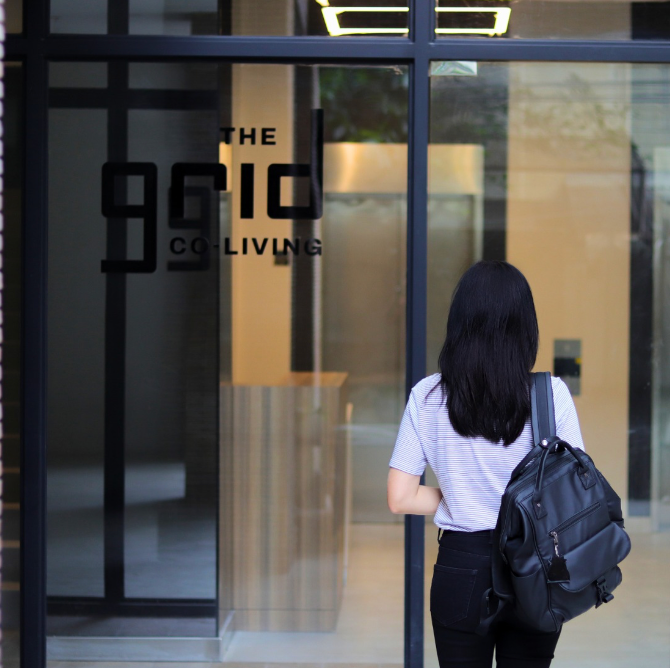 A person with dark hair wearing a backpack stands before a glass door labeled 