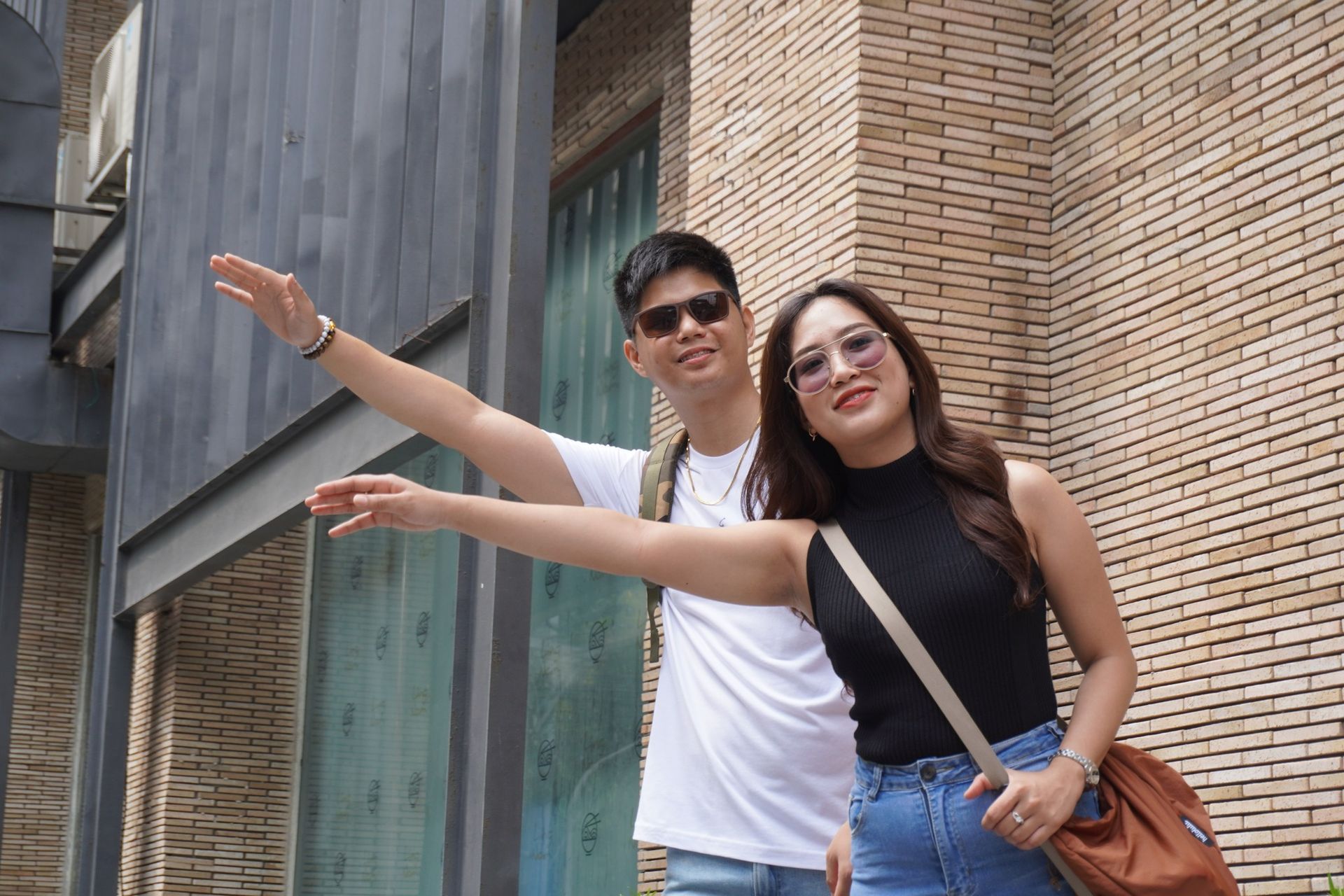 Two people, arms outstretched, hailing a taxi. Woman in black shirt, jeans; man in white shirt, sunglasses. Brick wall background.