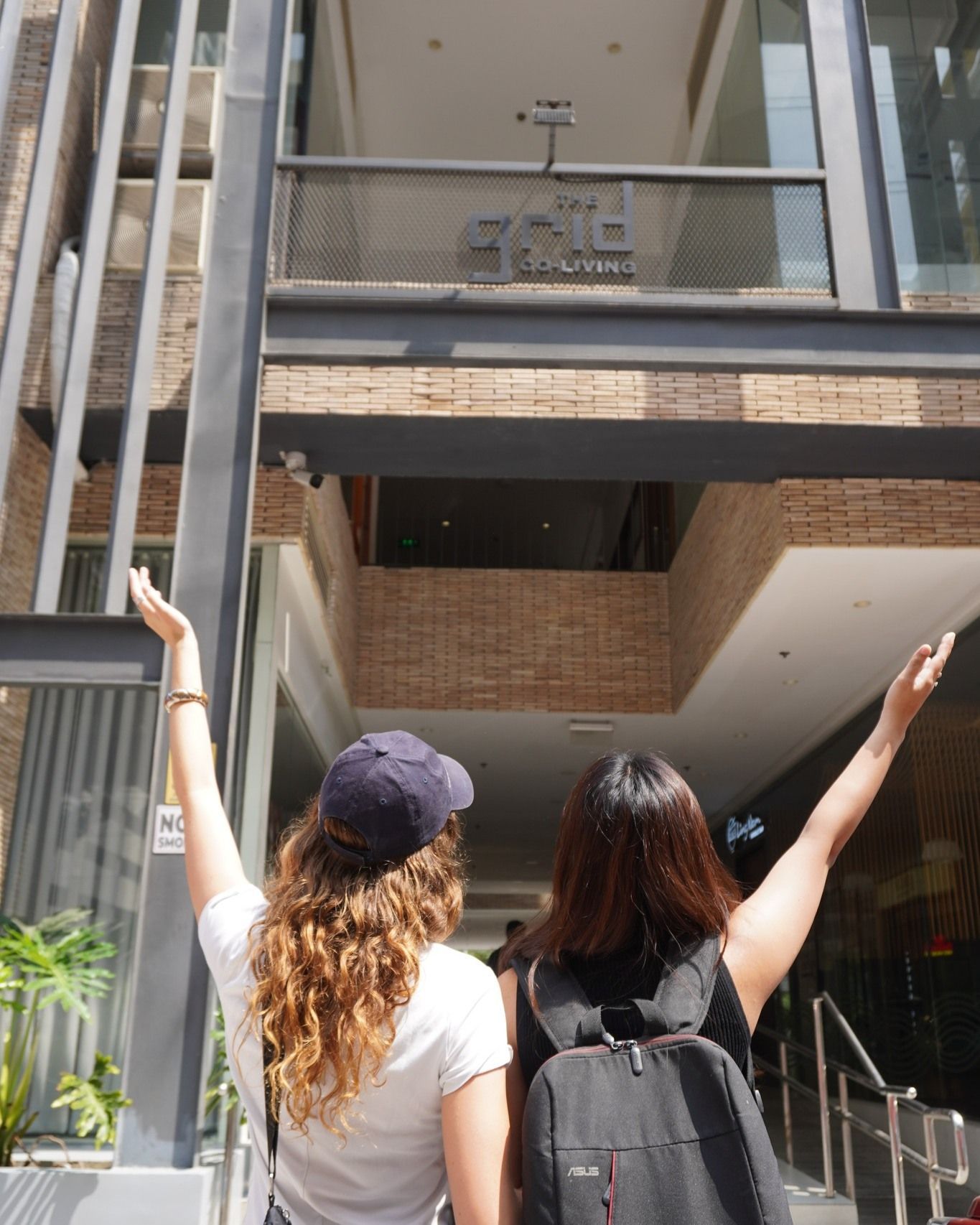 Two women with arms raised, standing in front of a building with a sign.