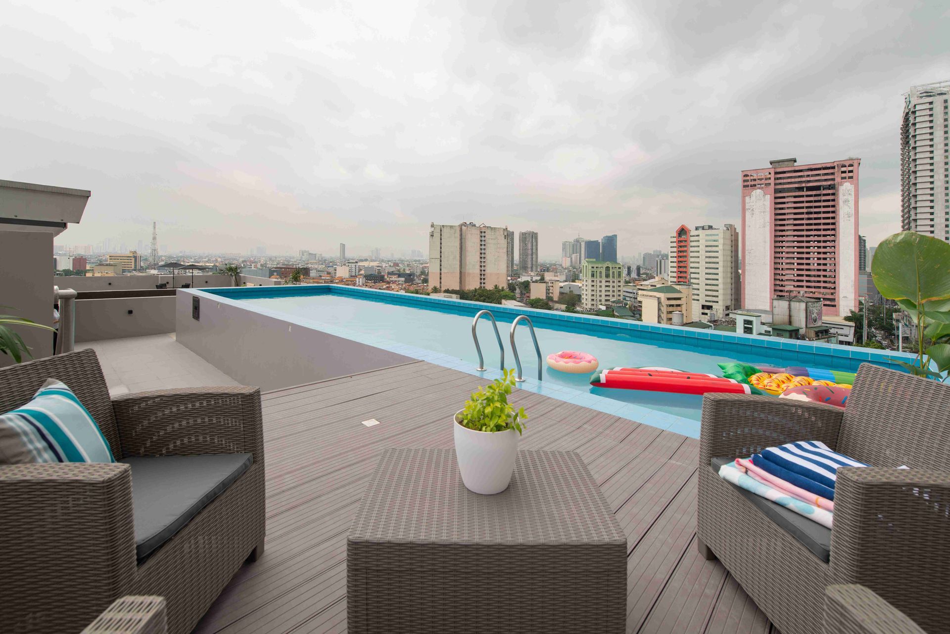 Rooftop pool with city skyline view. Wicker furniture on deck, pool floats in water. Overcast sky.