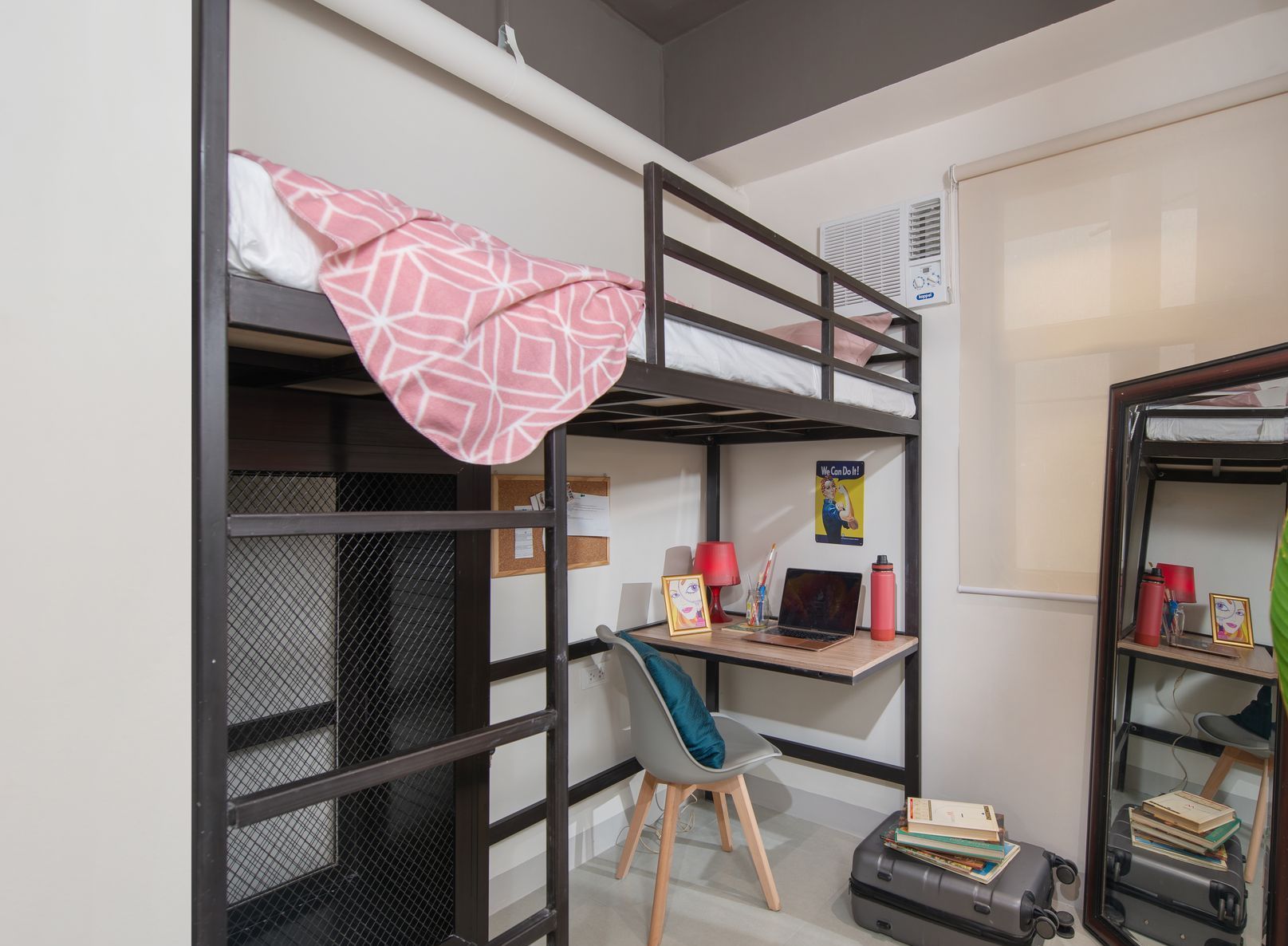 A small studio room with a black metal loft bed, a desk with a laptop, a chair, and a shelving unit.