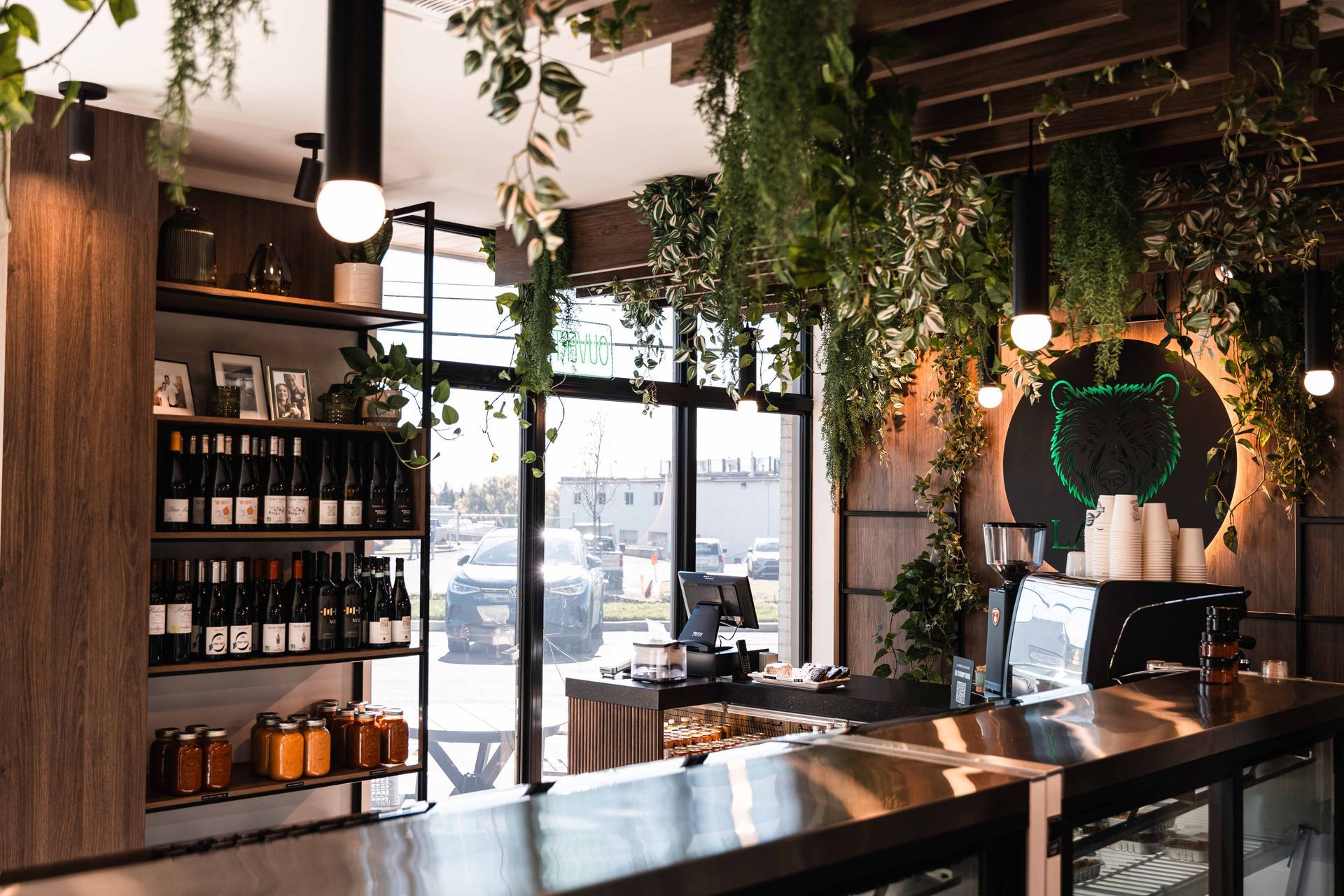 Intérieur d'un café avec un bar, des étagères de marchandises et des plantes suspendues près d'une grande fenêtre.
