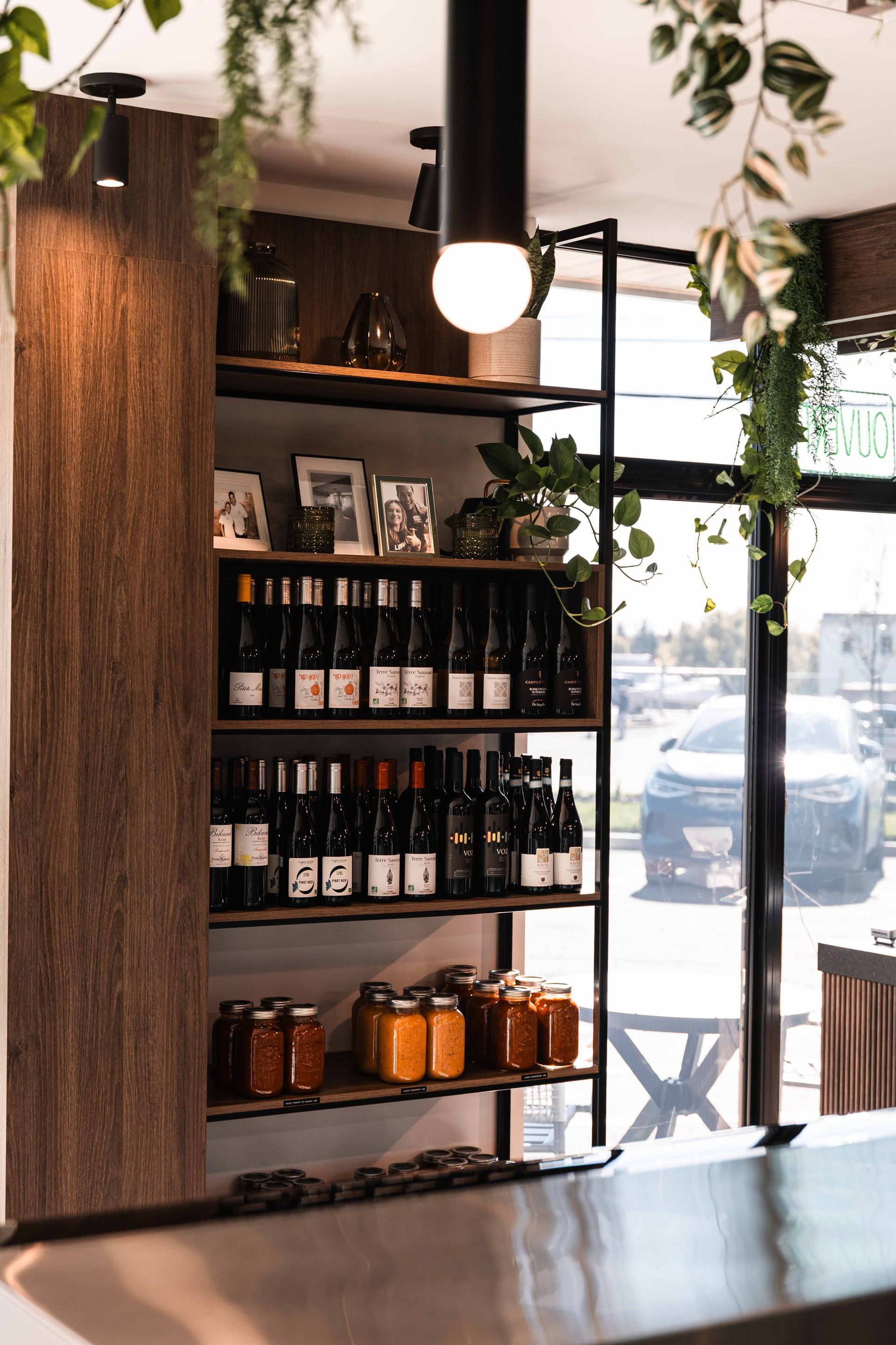 Étagère avec bouteilles de vin et bocaux de nourriture dans un magasin, avec une fenêtre et des plantes.