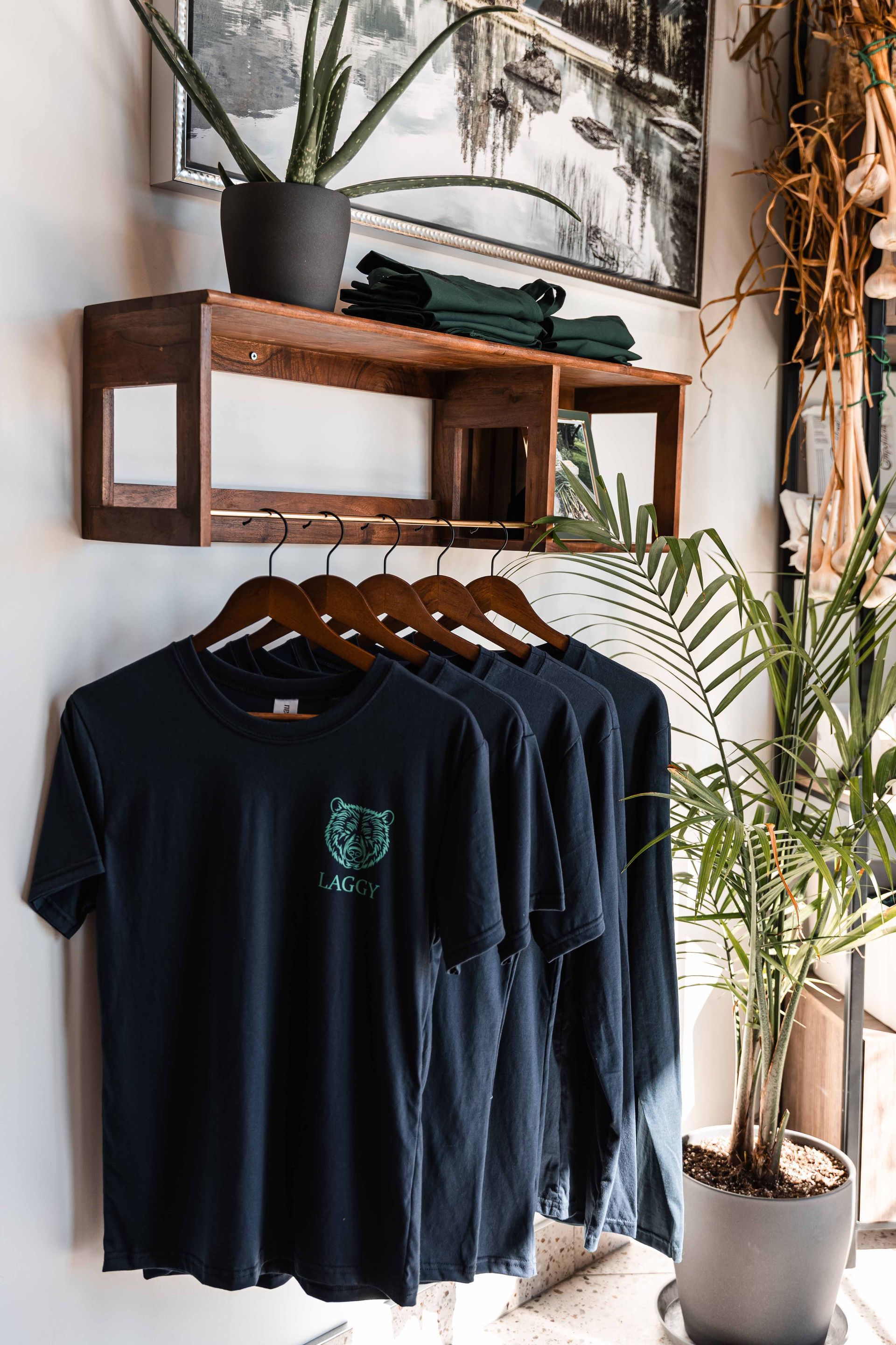 Des t-shirts bleu foncé sont suspendus à un portant sous une étagère en bois où se trouvent une plante et d'autres t-shirts.