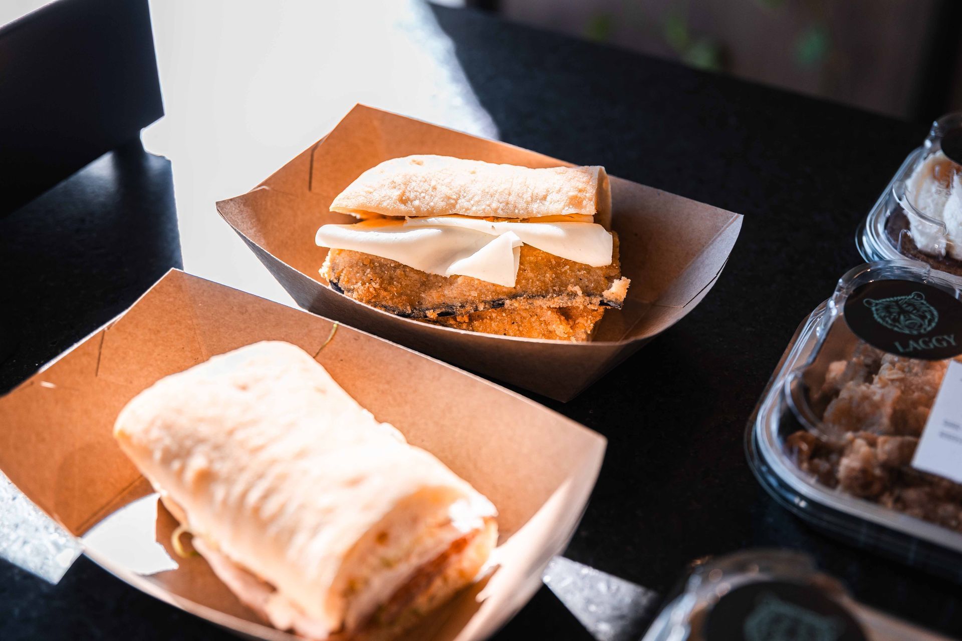 Deux sandwichs dans des boîtes en carton sur une table noire ; l'un ouvert avec du fromage, l'autre fermé.