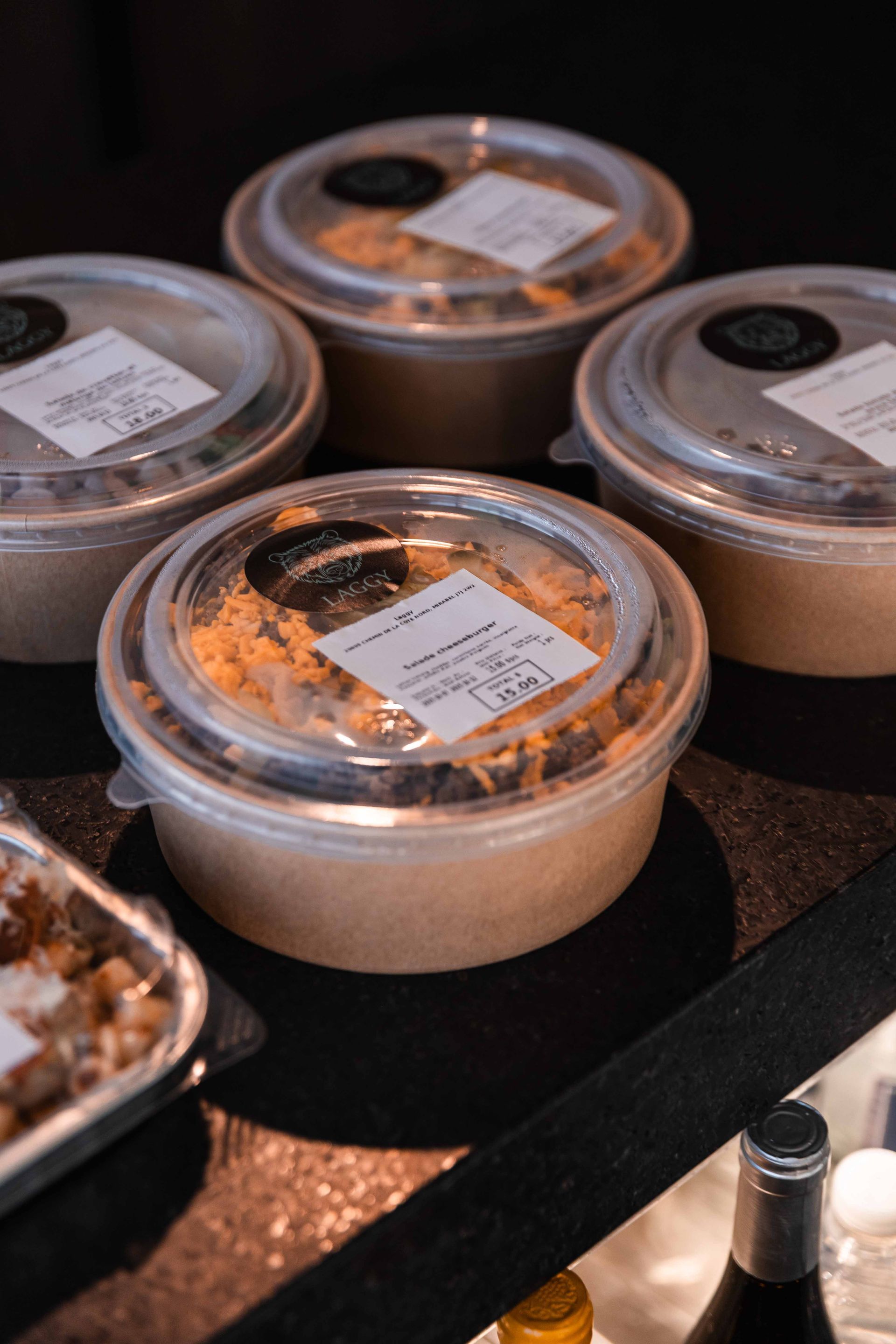Quatre récipients alimentaires sont posés sur une surface sombre, près d'un plateau contenant de la nourriture.