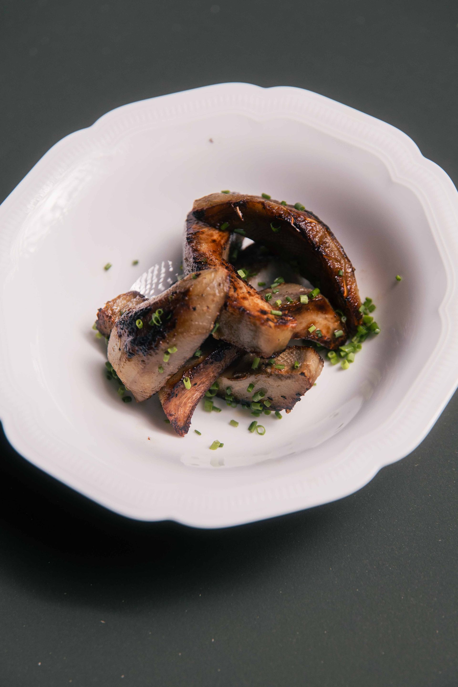 Tranches de poitrine de porc grillée aux ciboulettes dans un bol blanc, sur fond gris foncé.