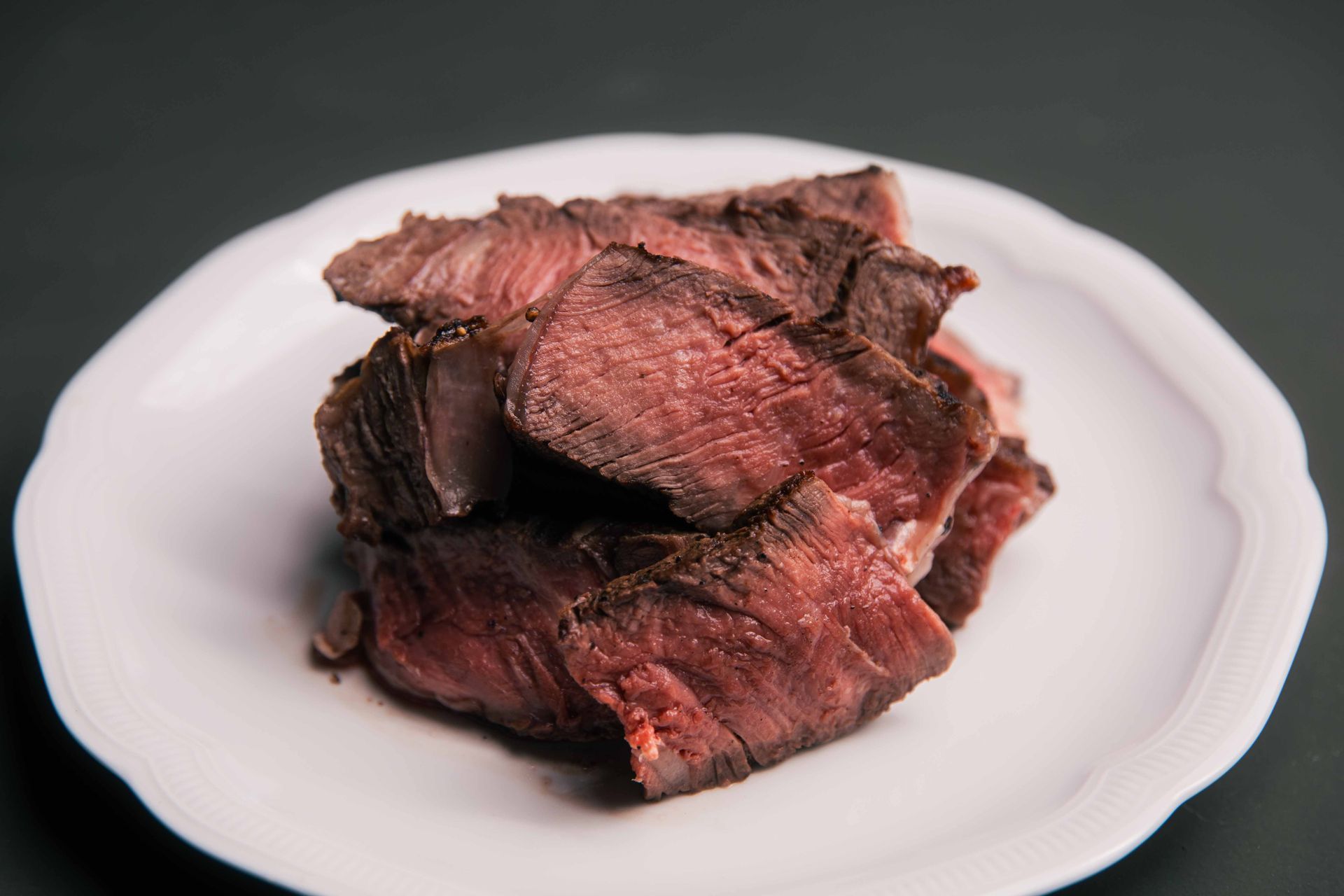 Tranches de steak cuit sur une assiette blanche sur fond sombre ; couleurs rouge et marron.