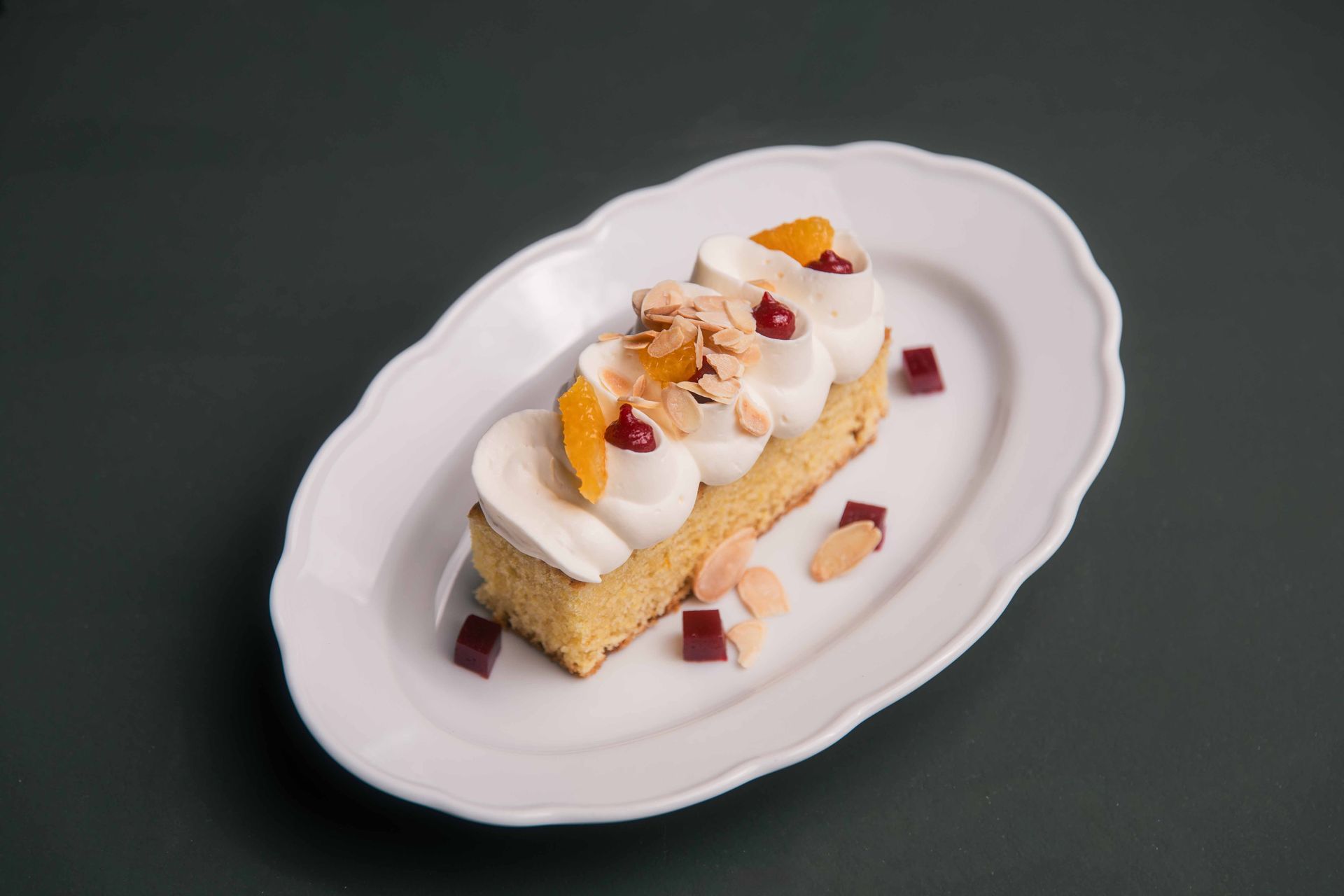 Une part de gâteau avec un glaçage blanc, des tranches d'orange et des amandes sur une assiette blanche.