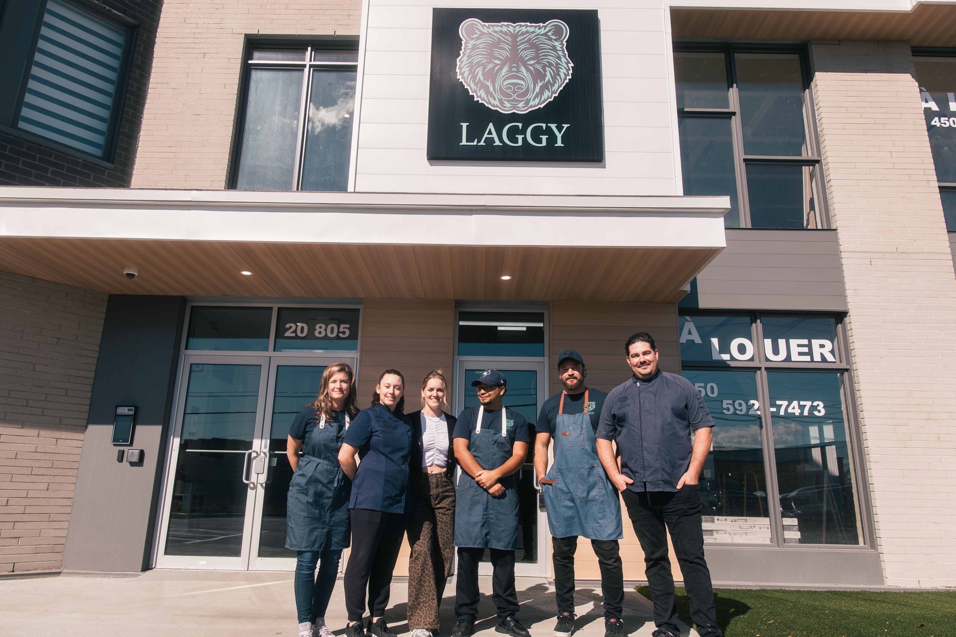 Un groupe de six personnes se tient devant un bâtiment portant l'enseigne « LAGGY ». Elles se trouvent devant une entrée.