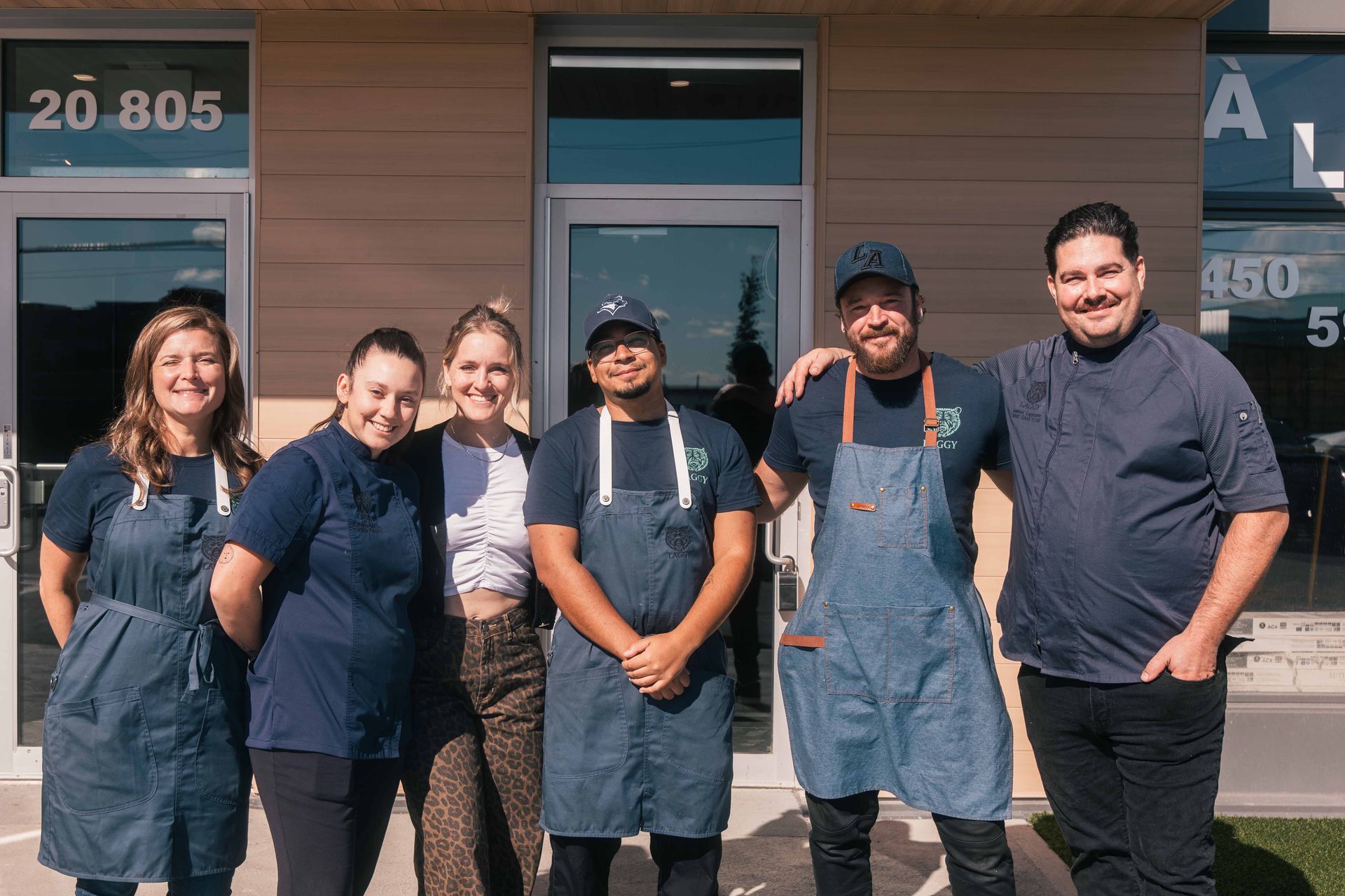 Six employés de restaurant posent devant un bâtiment, portant des tabliers et souriants.