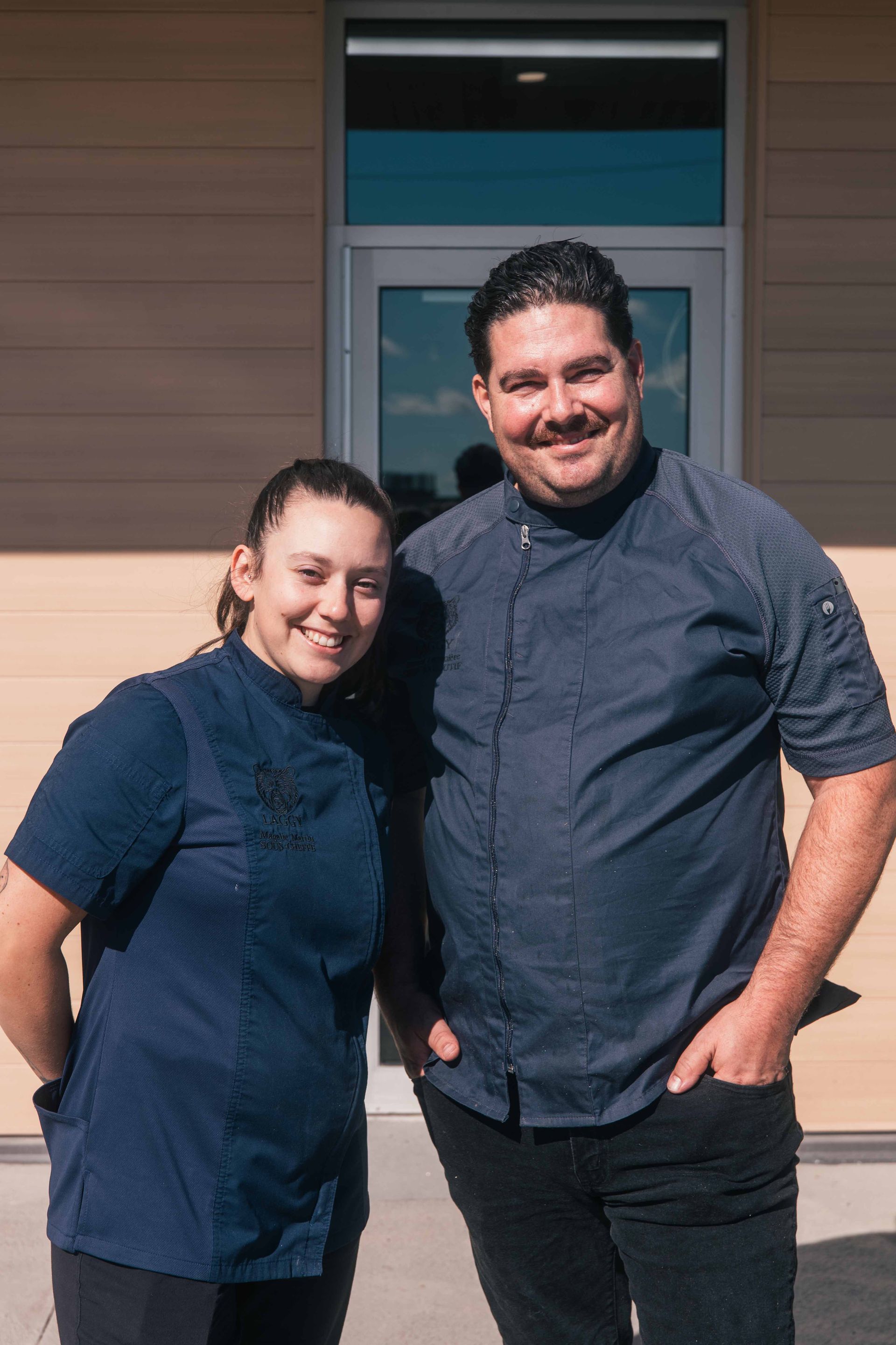 Deux chefs souriants posent devant un bâtiment. Tous deux portent des uniformes bleu foncé.