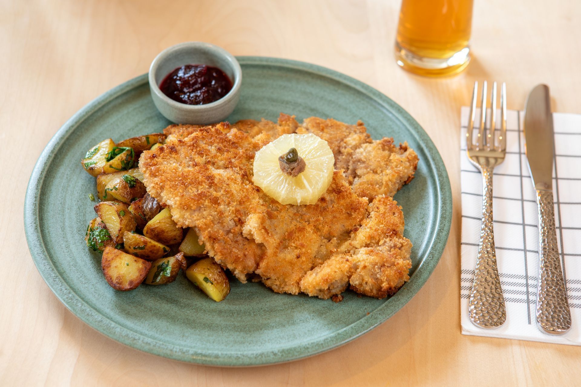 Paniertes Hähnchenschnitzel mit Kartoffeln, Beerensauce und einer Ananasscheibe auf einem türkisfarbenen Teller, dazu Besteck und Saft.