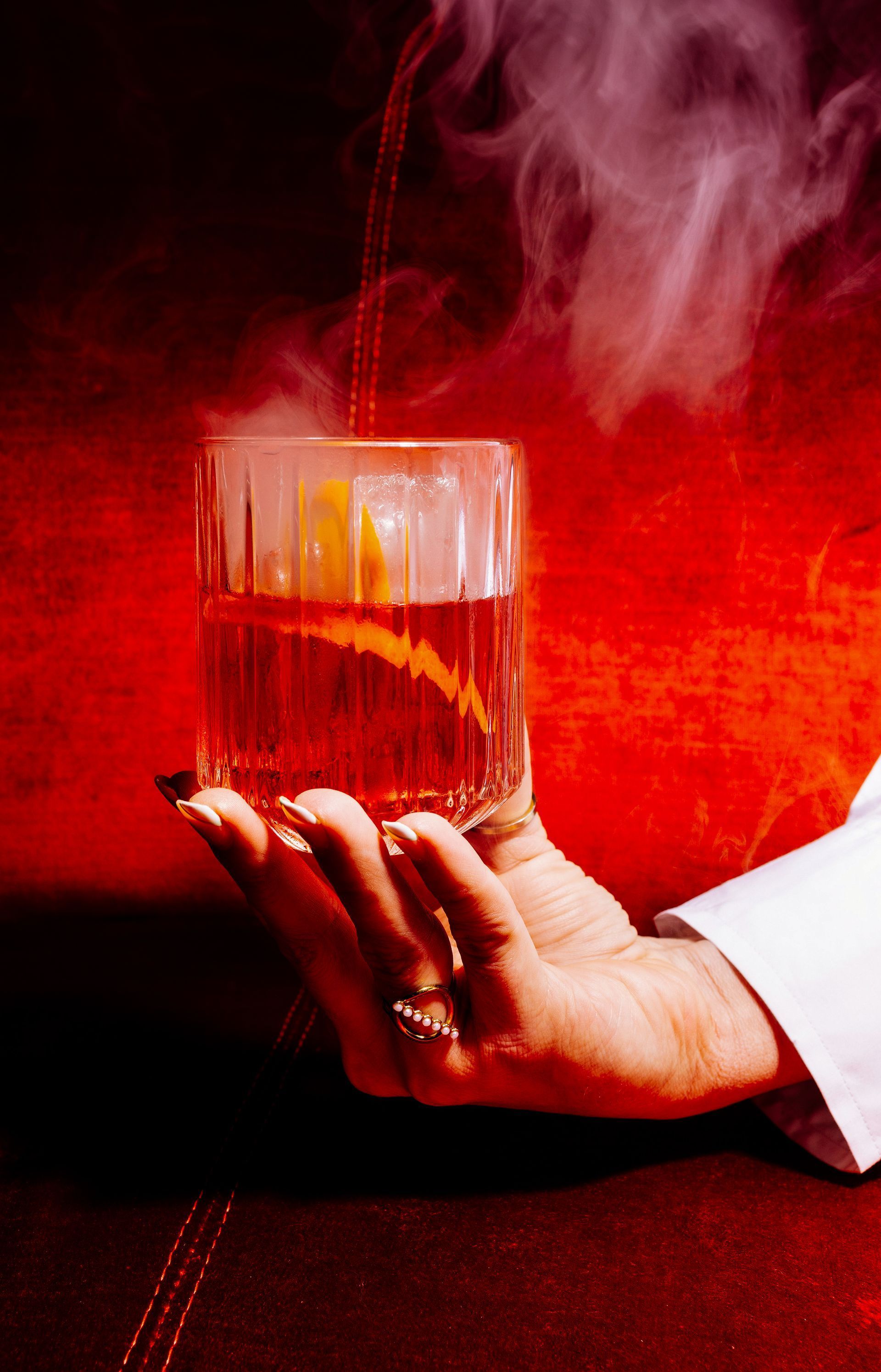 Eine Hand hält ein Glas bernsteinfarbenen Likörs, aus dem Rauch aufsteigt, vor einem leuchtend roten Hintergrund.