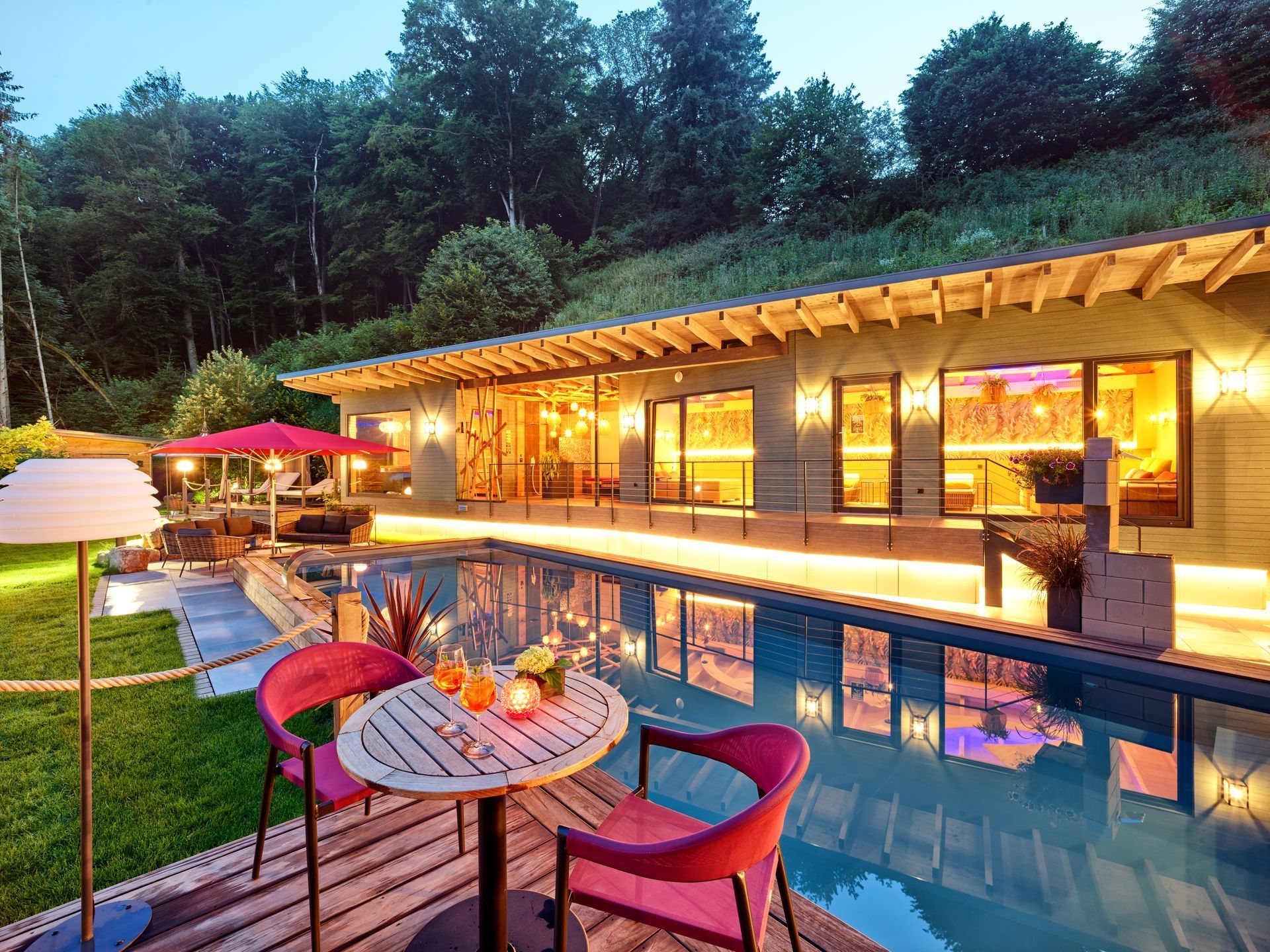 Luxuriöse Poolvilla in der Abenddämmerung mit beleuchteter Terrasse, roten Gartenstühlen und einem spiegelnden Swimmingpool