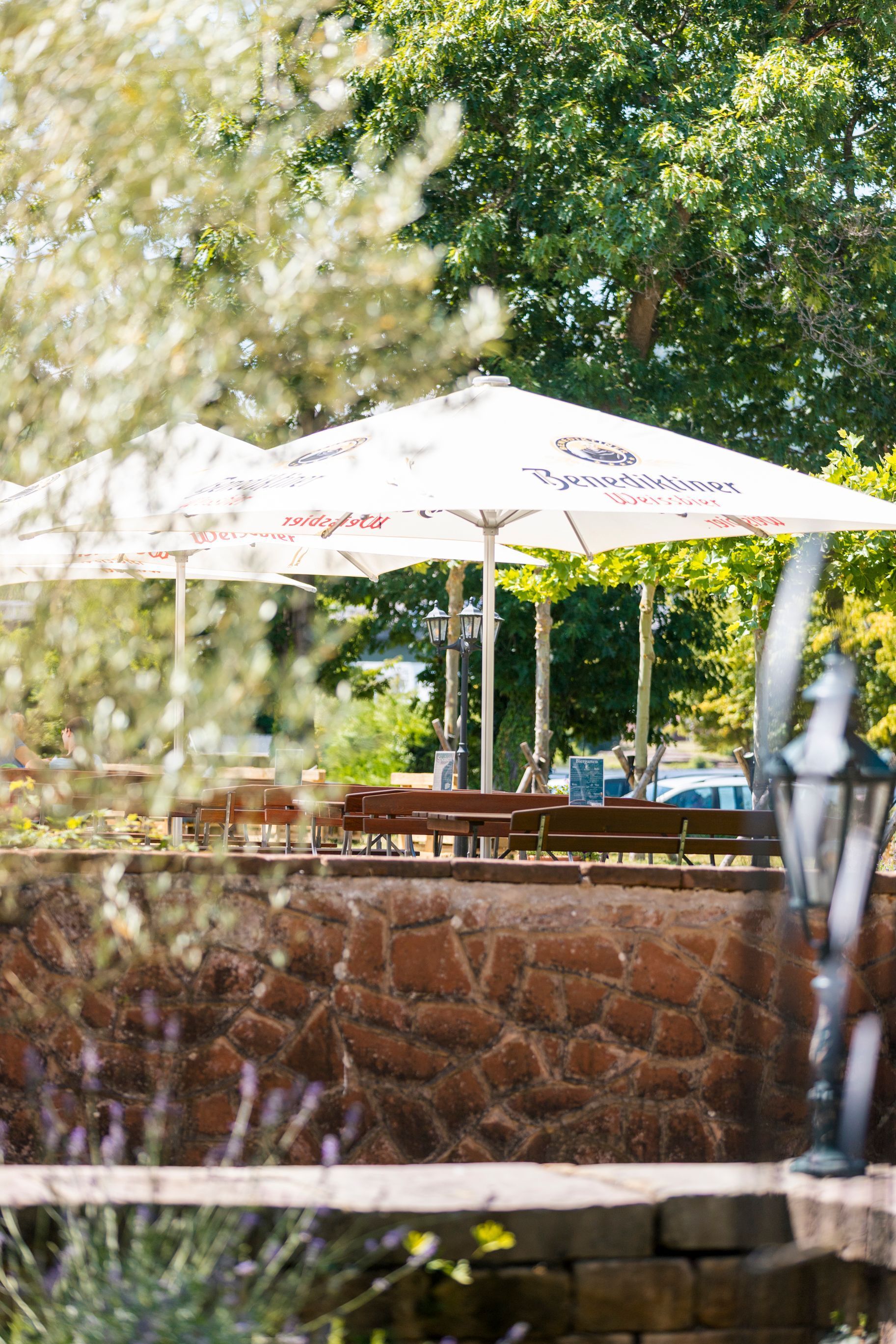 Terrasse im Freien mit weißen Sonnenschirmen hinter einer Steinmauer, umgeben von Bäumen und Grün.