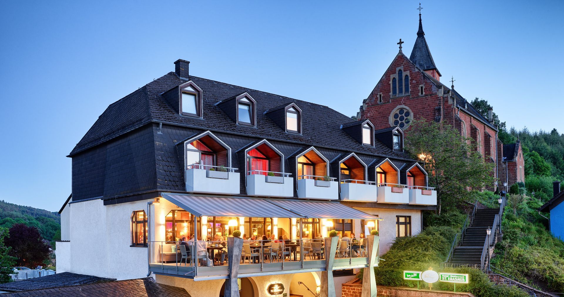 Historisches weißes Gasthaus mit in der Abenddämmerung erleuchteten Fenstern, neben einer roten Backsteinkirche und Grünanlagen
