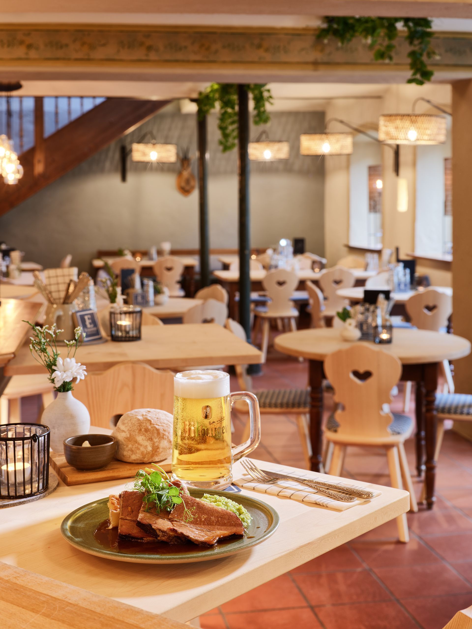 Gemütlicher Café-Essbereich mit einem angerichteten Menü und Bier im Vordergrund, warmen Pendelleuchten und leeren Tischen.