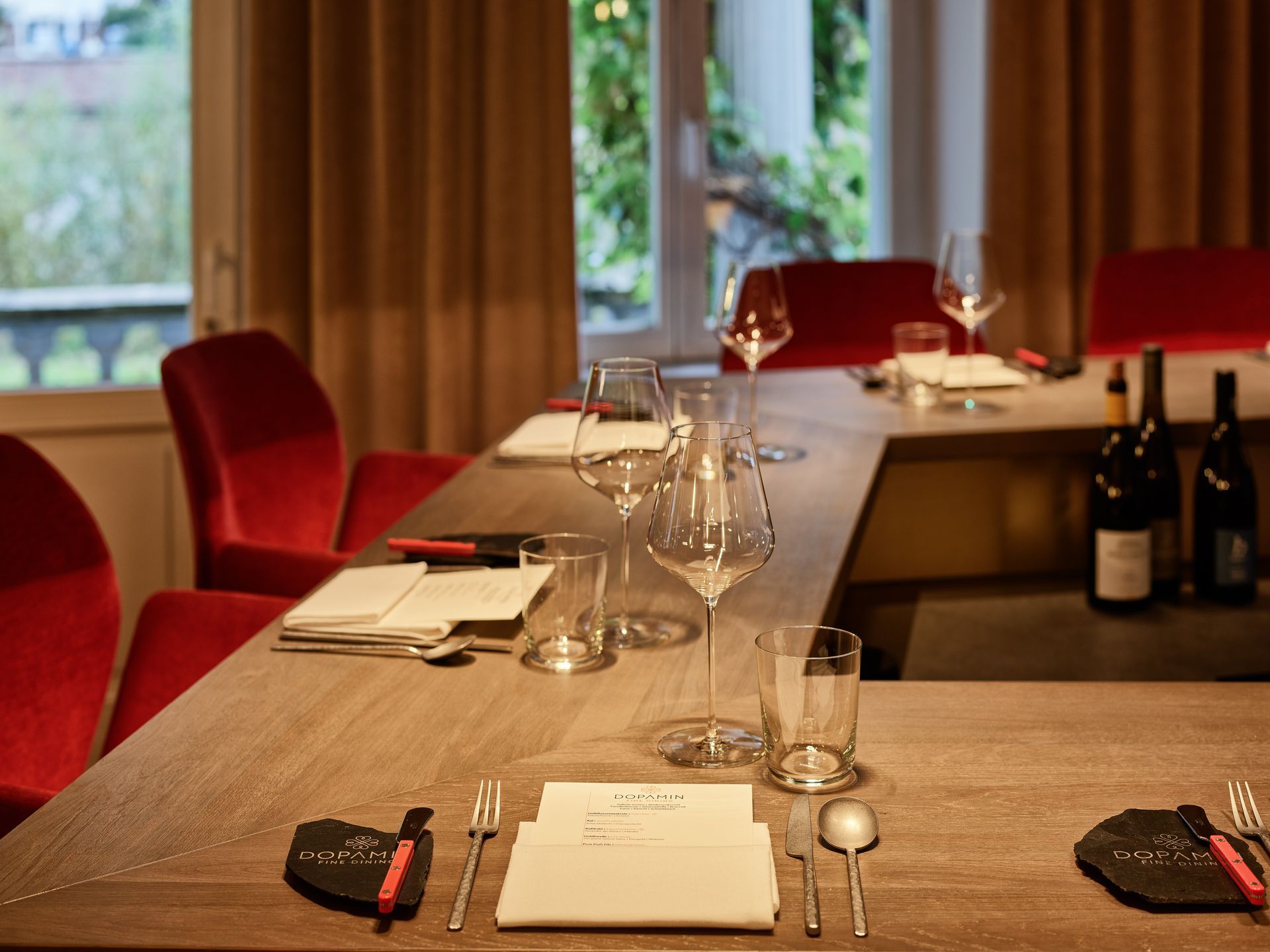 Ein ordentlich gedeckter Restauranttisch mit roten Stühlen, Weinflaschen, Gläsern und Servietten steht in der Nähe eines Fensters.
