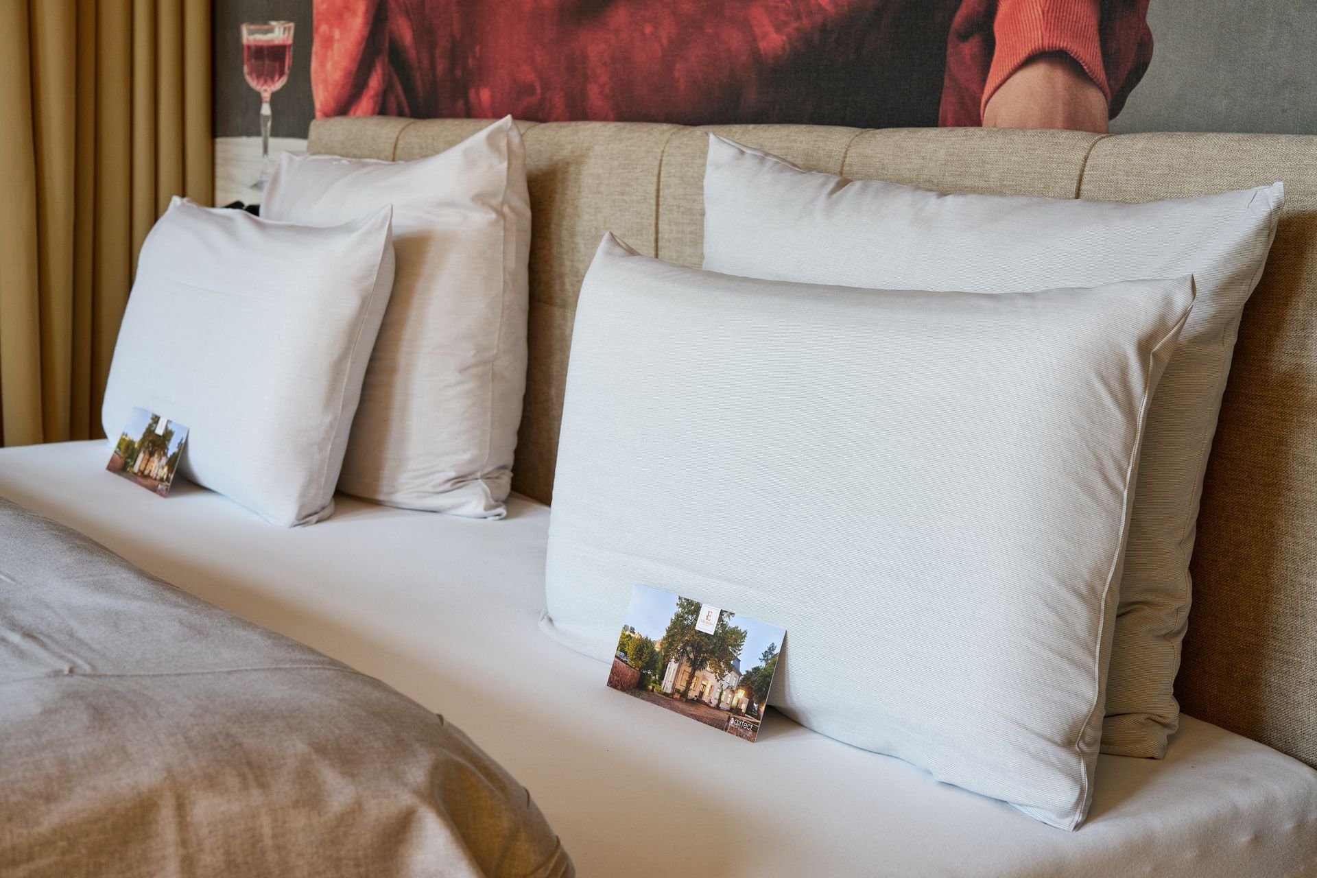 Hotelbett mit weißen Kissen, beiger Decke und kleinen Dekoartikeln auf dem Ablagefach am Kopfteil.