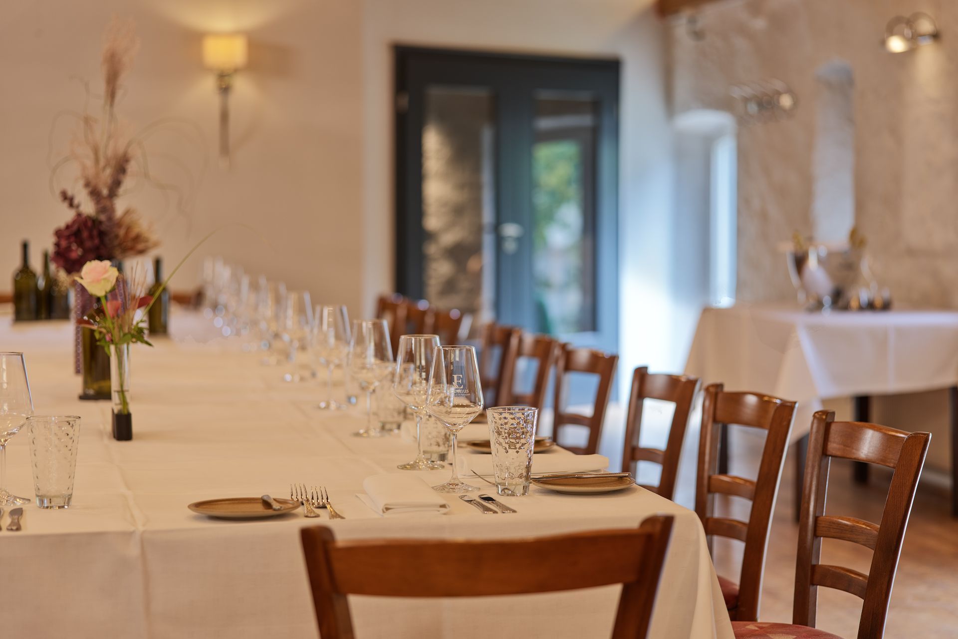Eine lange Festtafel, gedeckt mit weißen Tischdecken und Weingläsern, in einem sanft beleuchteten Speisesaal.