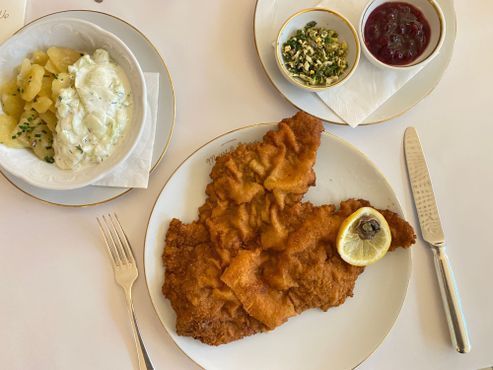 Ein Teller mit paniertem, gebratenem Fisch und Zitrone, serviert mit Kartoffeln, Soßen und Besteck auf einem Tisch.