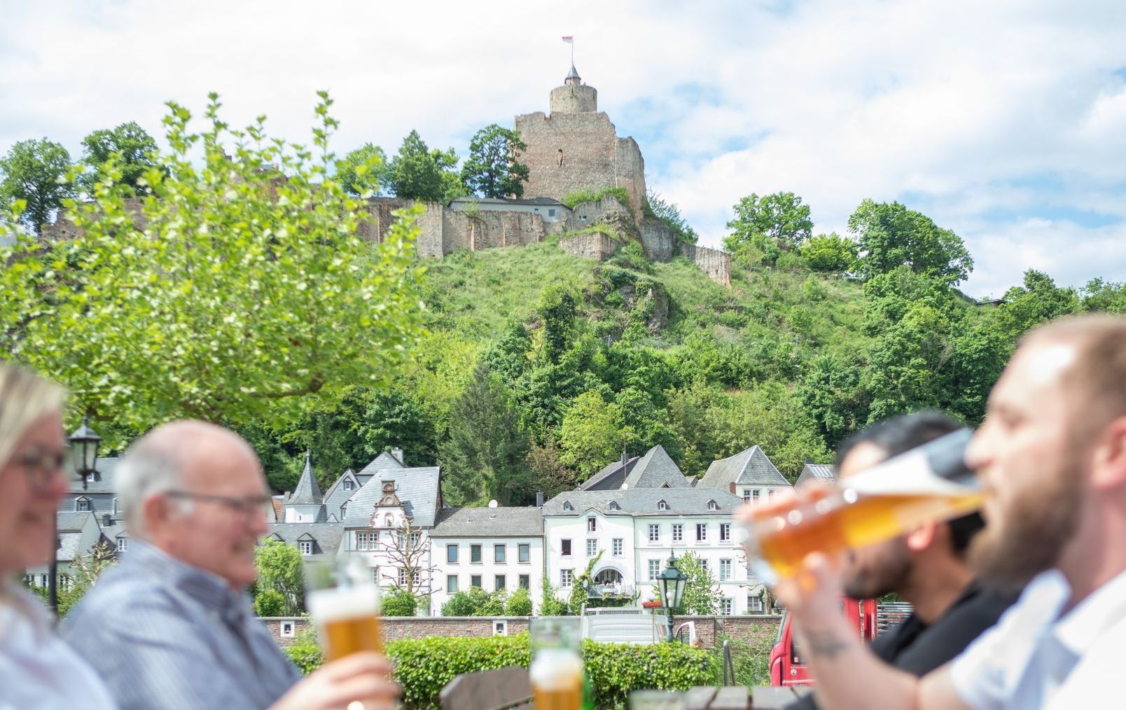 Menschen stoßen vor Burgen am Hang und Dorfhäusern mit Biergläsern an.