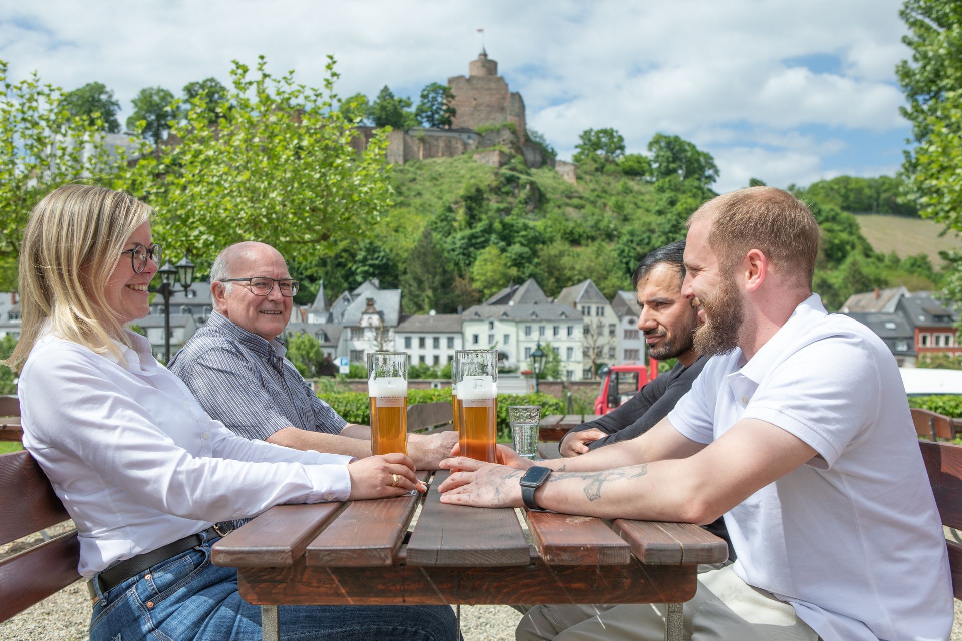 Menschen unterhalten sich bei einem Bier an einem Tisch im Freien, im Hintergrund erhebt sich eine Burg auf einem grünen Hügel.