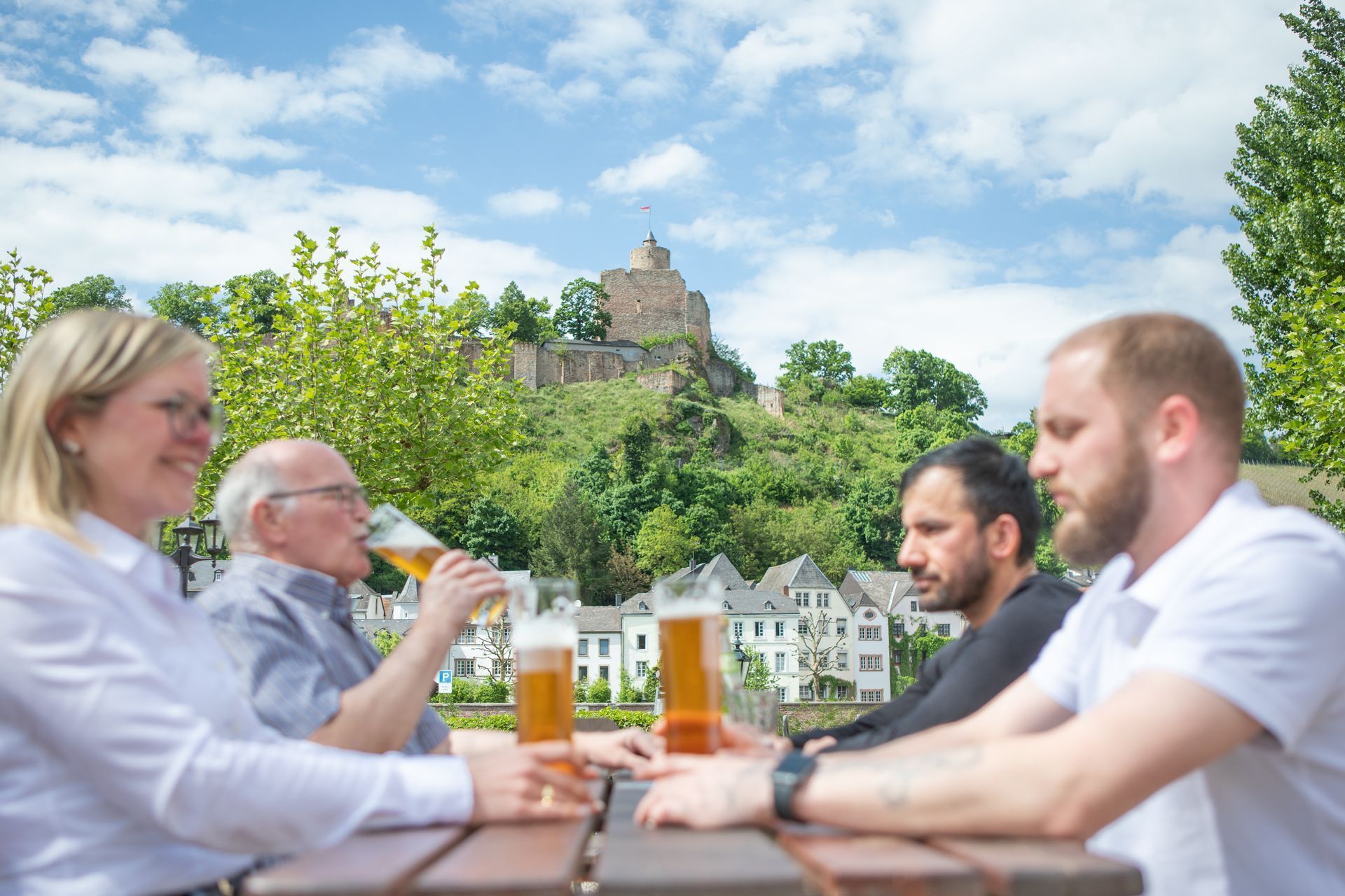 Menschen sitzen an einem Picknicktisch und trinken Bier im Freien; im Hintergrund erhebt sich eine Burg auf dem Hügel.