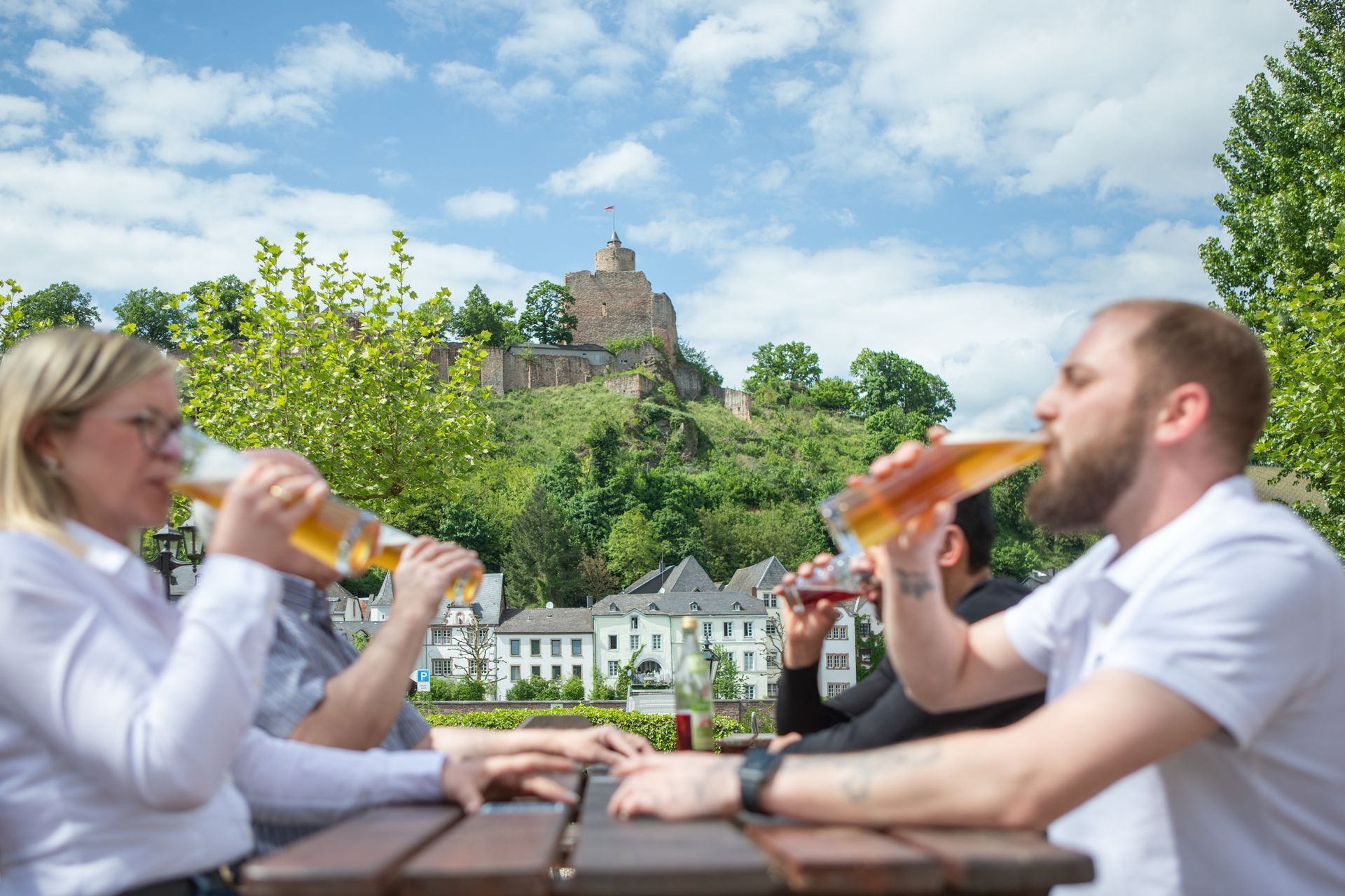 Zwei Personen trinken Bier an einem Tisch im Freien; im Hintergrund ist eine Burg auf einem Hügel zu sehen.