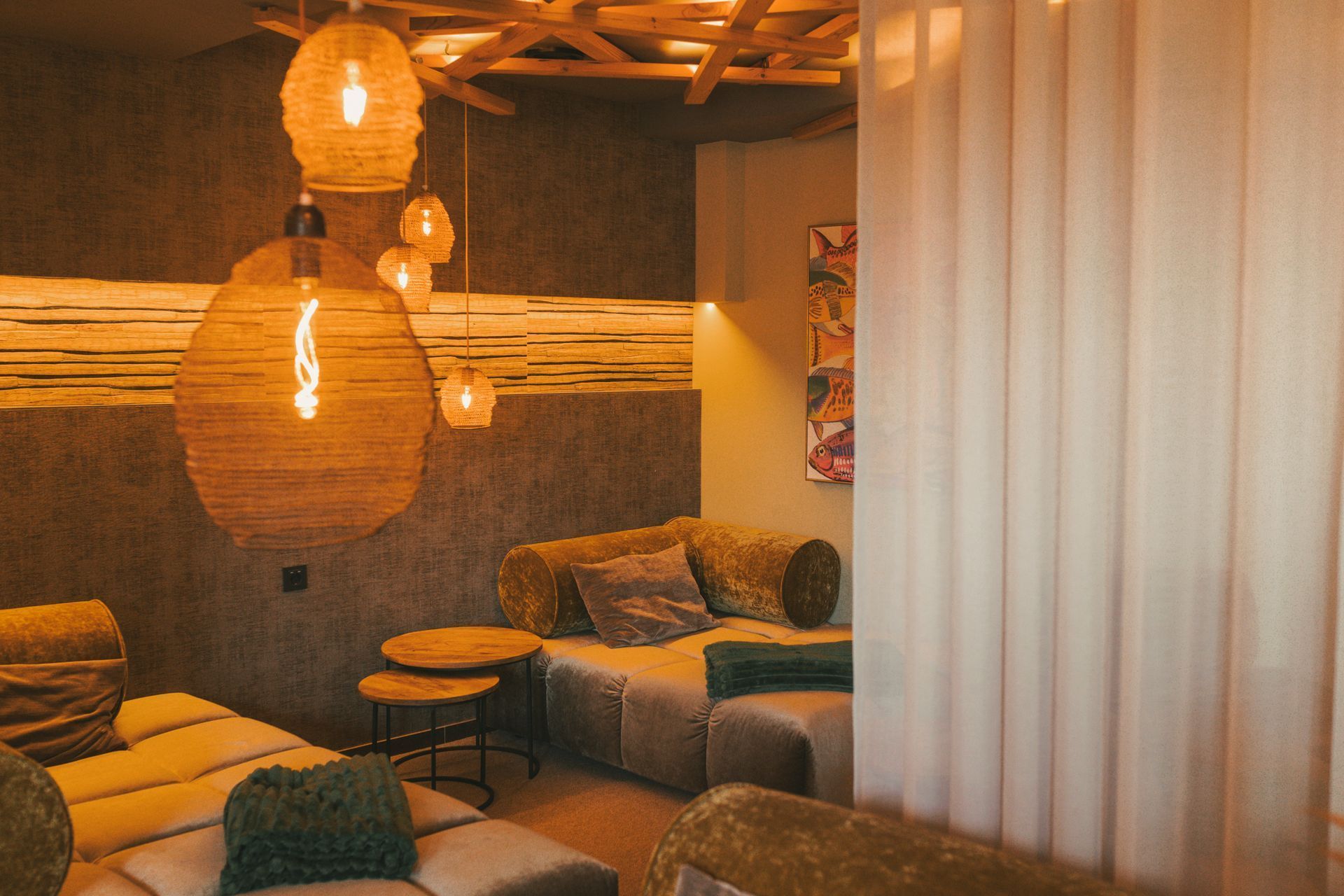 Gemütliche Lounge mit gewebten Pendelleuchten, grauen Sofas und warmem, bernsteinfarbenem Licht hinter einem Vorhang.