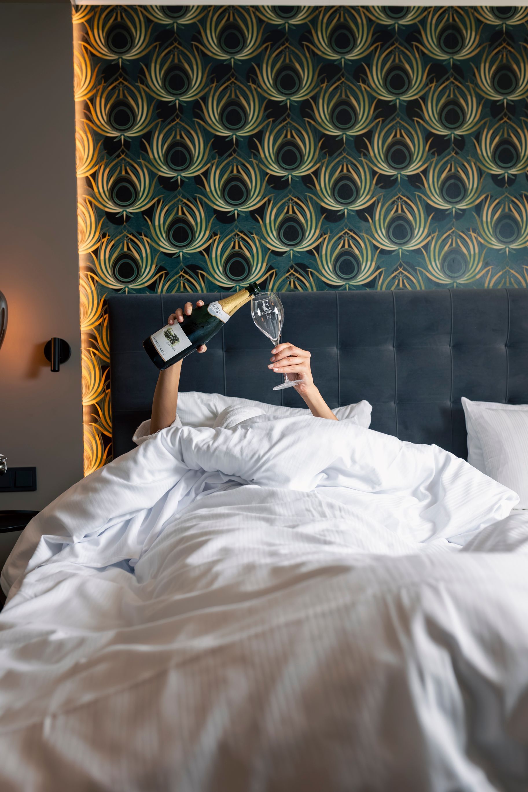 Eine Person liegt im Bett und hält ein Weinglas und eine Flasche; im Hintergrund ist eine auffällig gemusterte Tapete zu sehen.