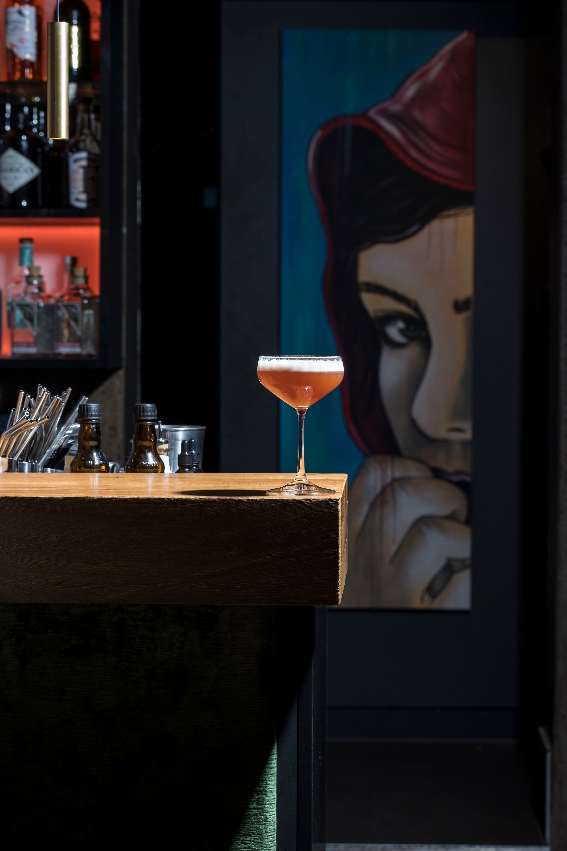 Ein Cocktail auf einer hölzernen Bartheke vor einem großen gemalten Porträt in einer schwach beleuchteten Bar.